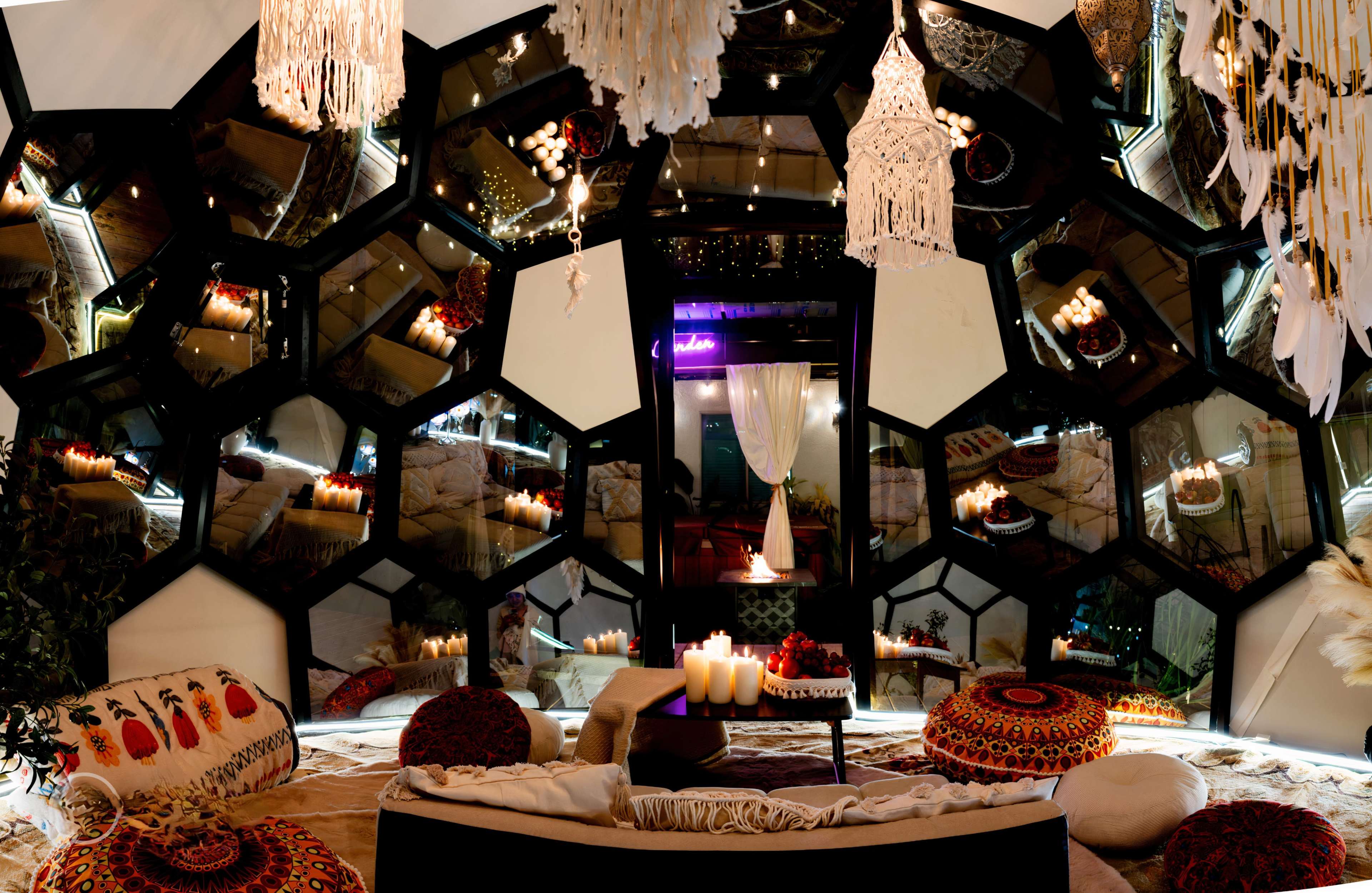 The image depicts a cozy lounge area adorned with hexagonal mirrors, candles, and decorative cushions arranged on a plush floor.
