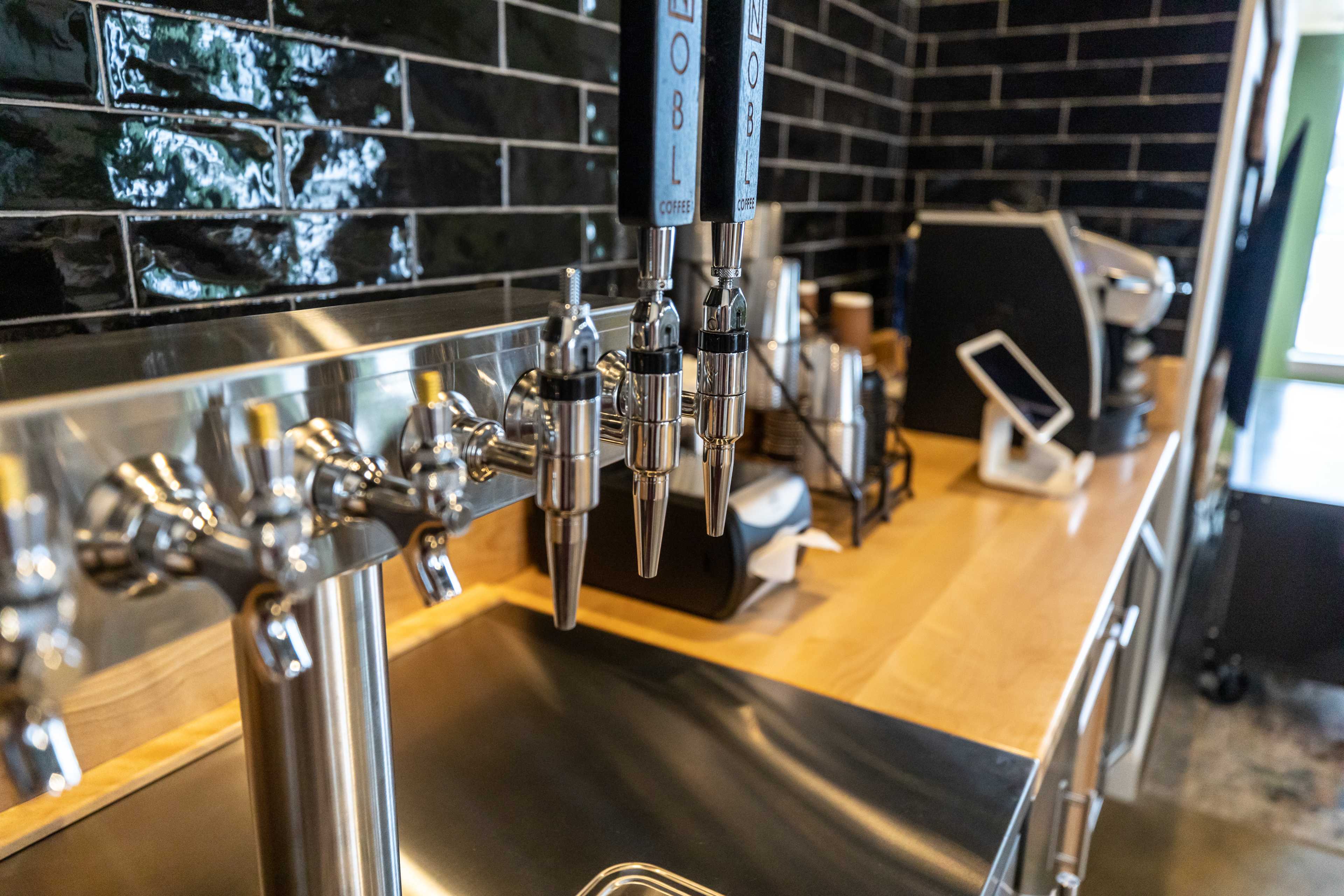 The image shows a row of four metal beverage taps next to a countertop with a coffee machine and various supplies in a kitchen setting.