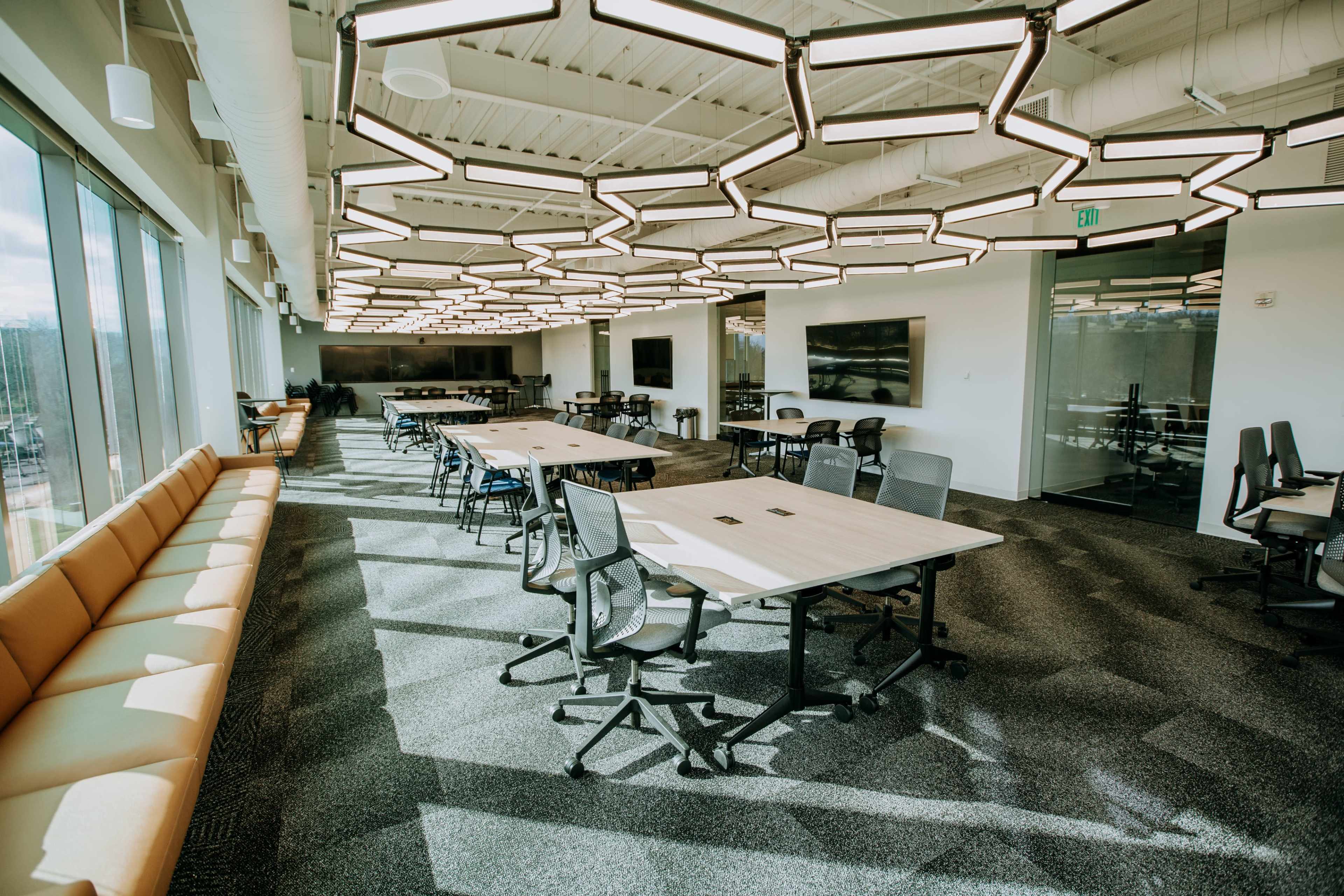 The image shows a modern office space featuring multiple tables and chairs, large windows with natural light, and a unique hexagonal light fixture overhead.