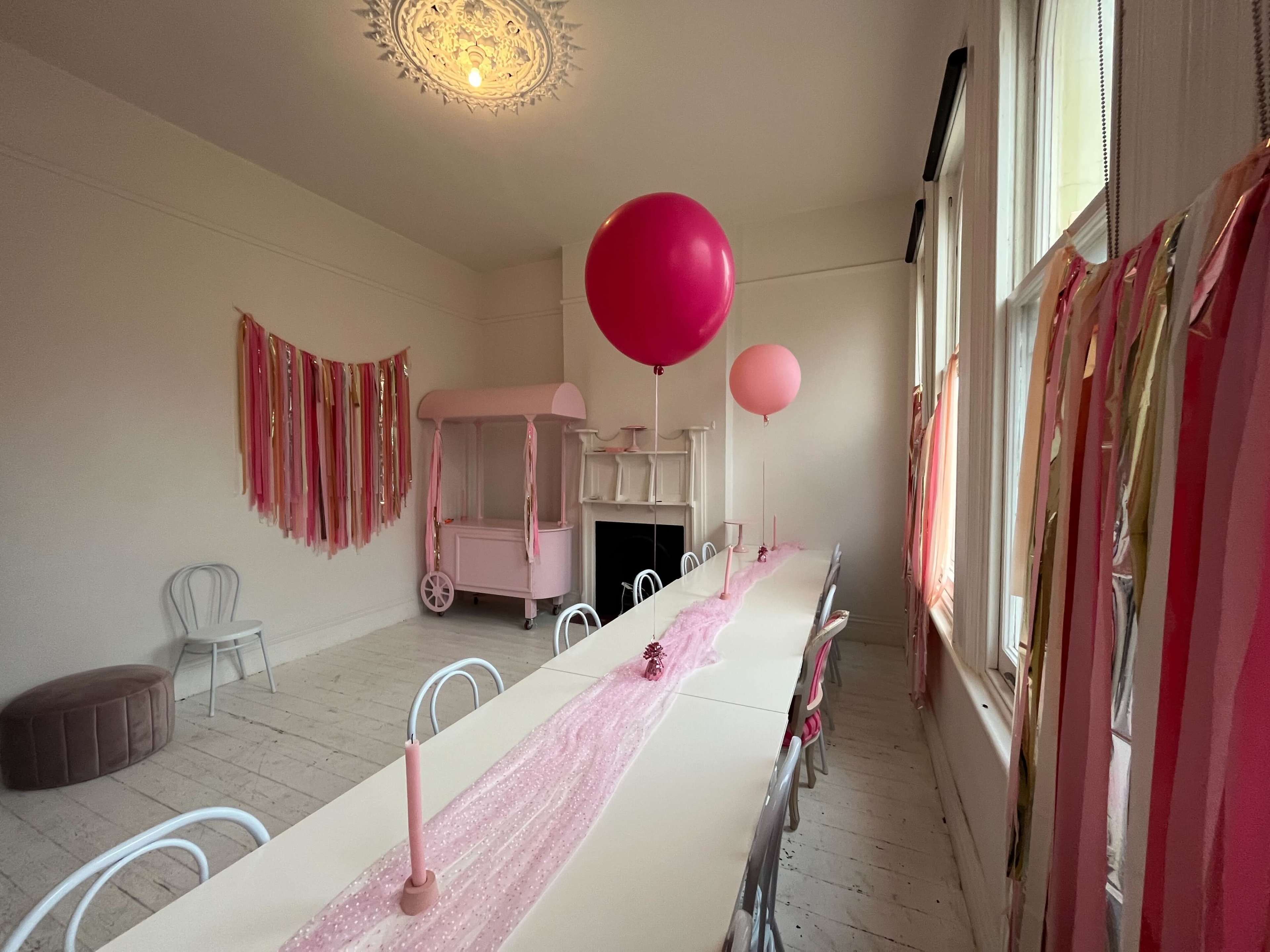 A long table is set with a pink tablecloth and candles, flanked by colorful streamers and two large pink balloons in a bright room.