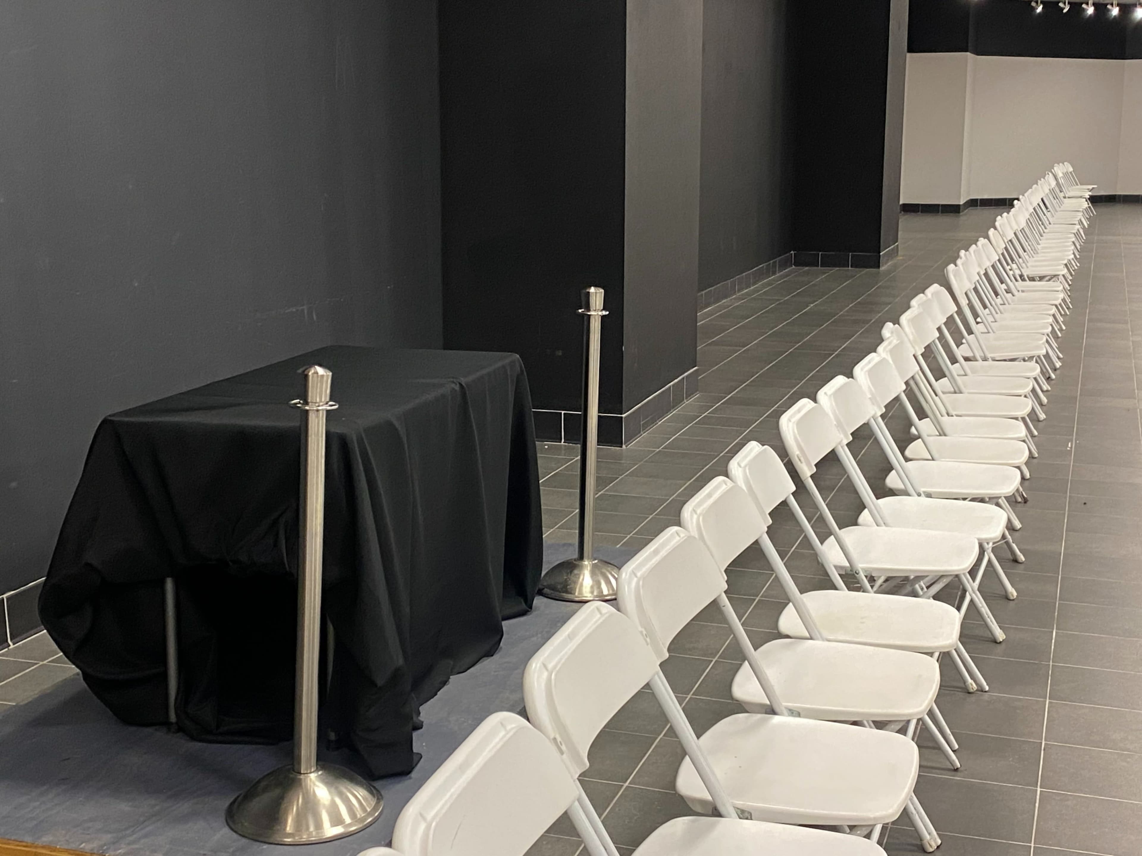 A row of white folding chairs is arranged neatly in a hallway next to a table covered with a black cloth.