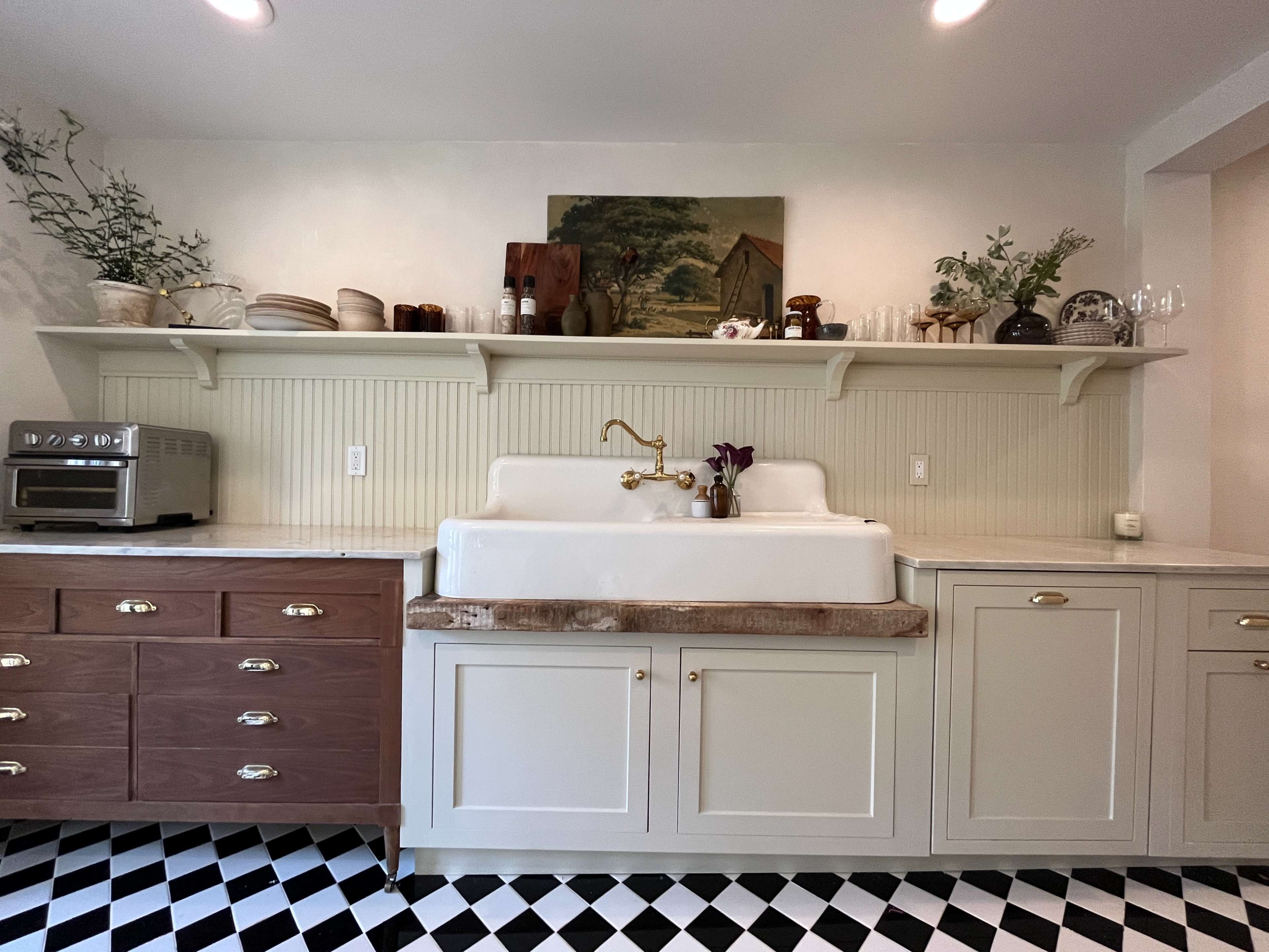 The image shows a kitchen featuring a large farmhouse sink with a gold faucet, wooden shelves displaying various kitchen items, and a checkered black-and-white floor.