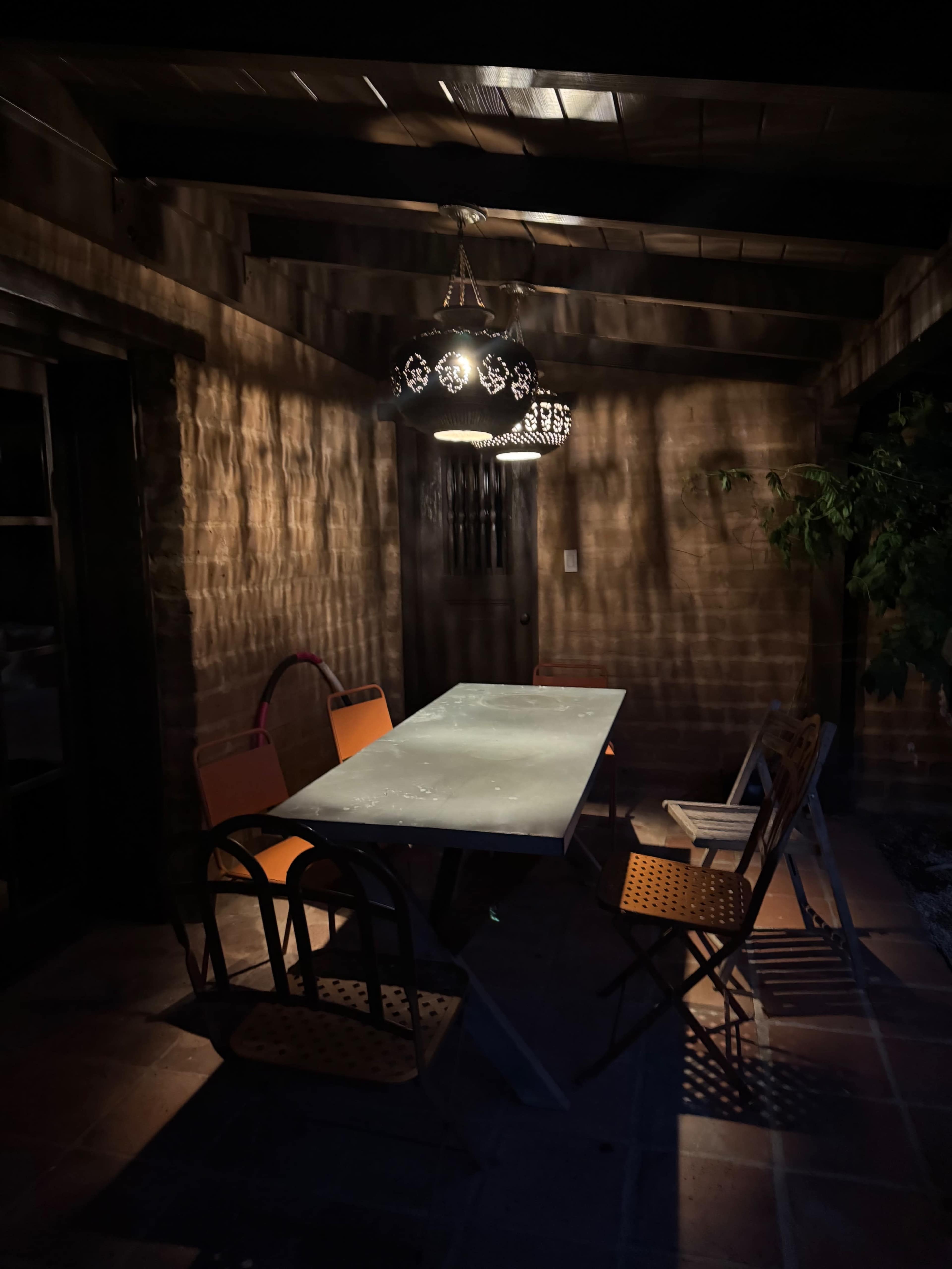 A rectangular table with several chairs is placed under a hanging light in a dimly lit outdoor area.