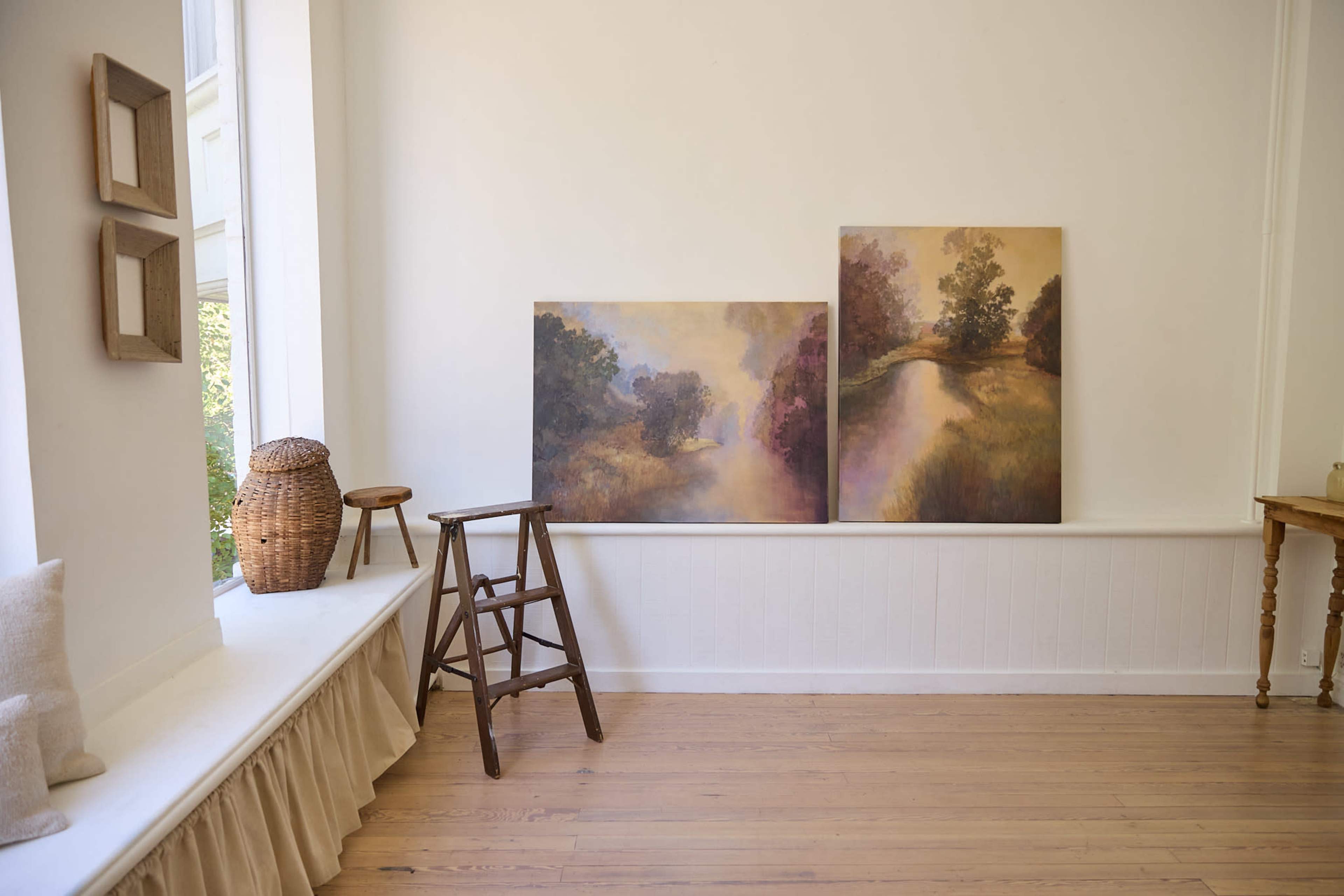 The image shows an airy, minimalist room featuring two large paintings on the wall, a wooden stool, a wicker basket, and a window with a ledge.