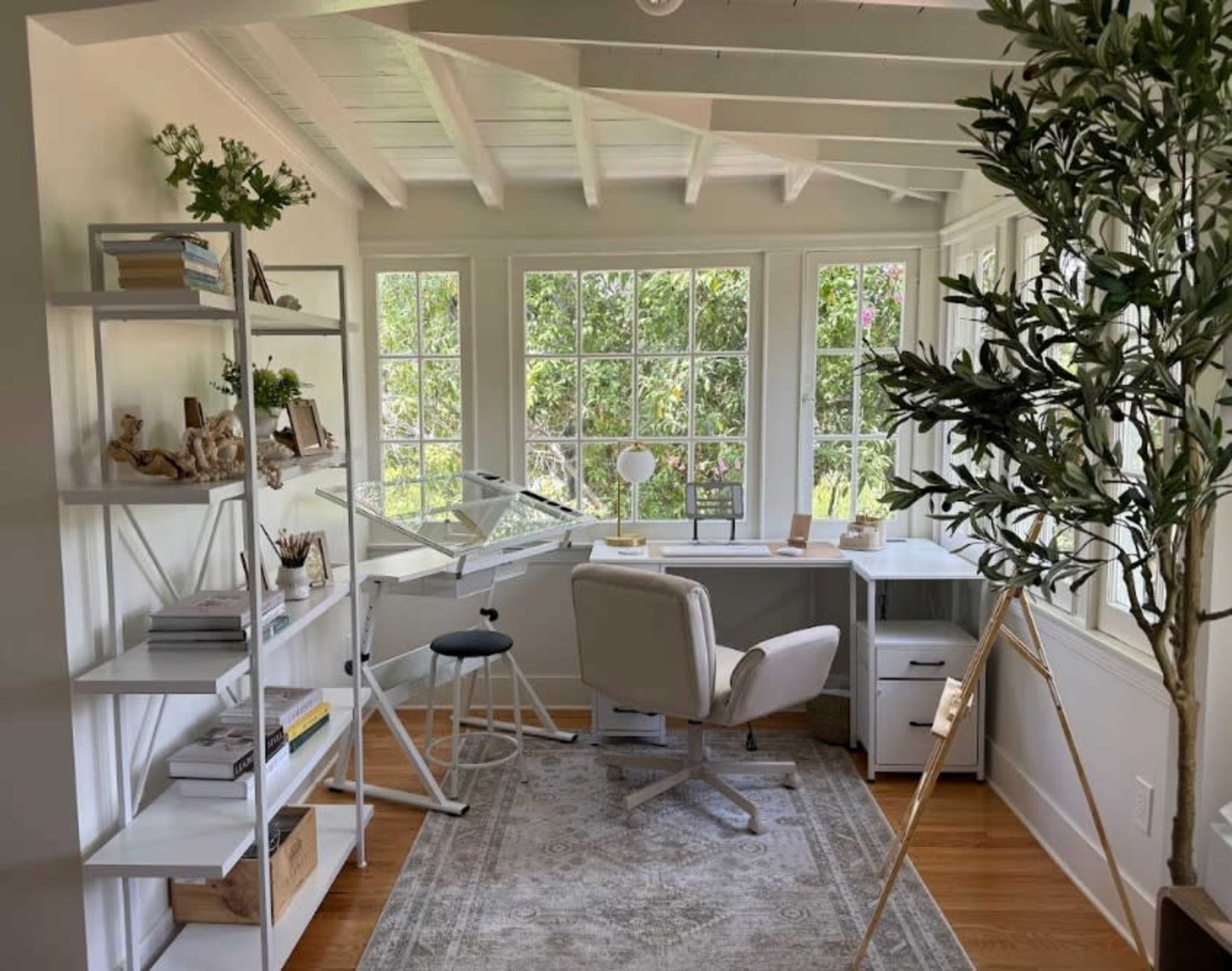 The image shows a bright and airy home office with a drafting table, a comfortable chair, a desk, and large windows overlooking greenery outside.