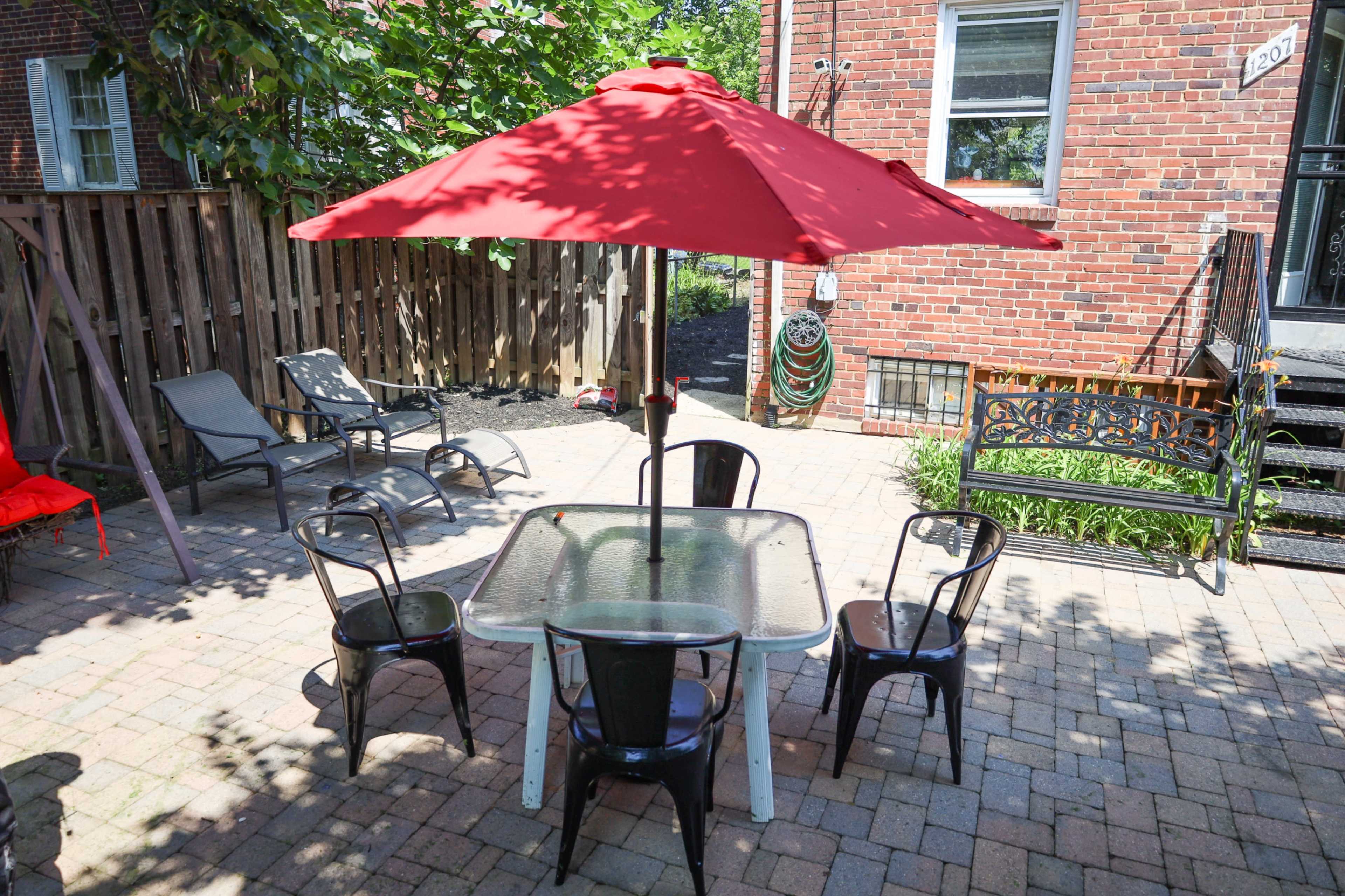 The image shows a patio area with a glass table, four black chairs, and a large red umbrella, surrounded by brick walls and greenery.