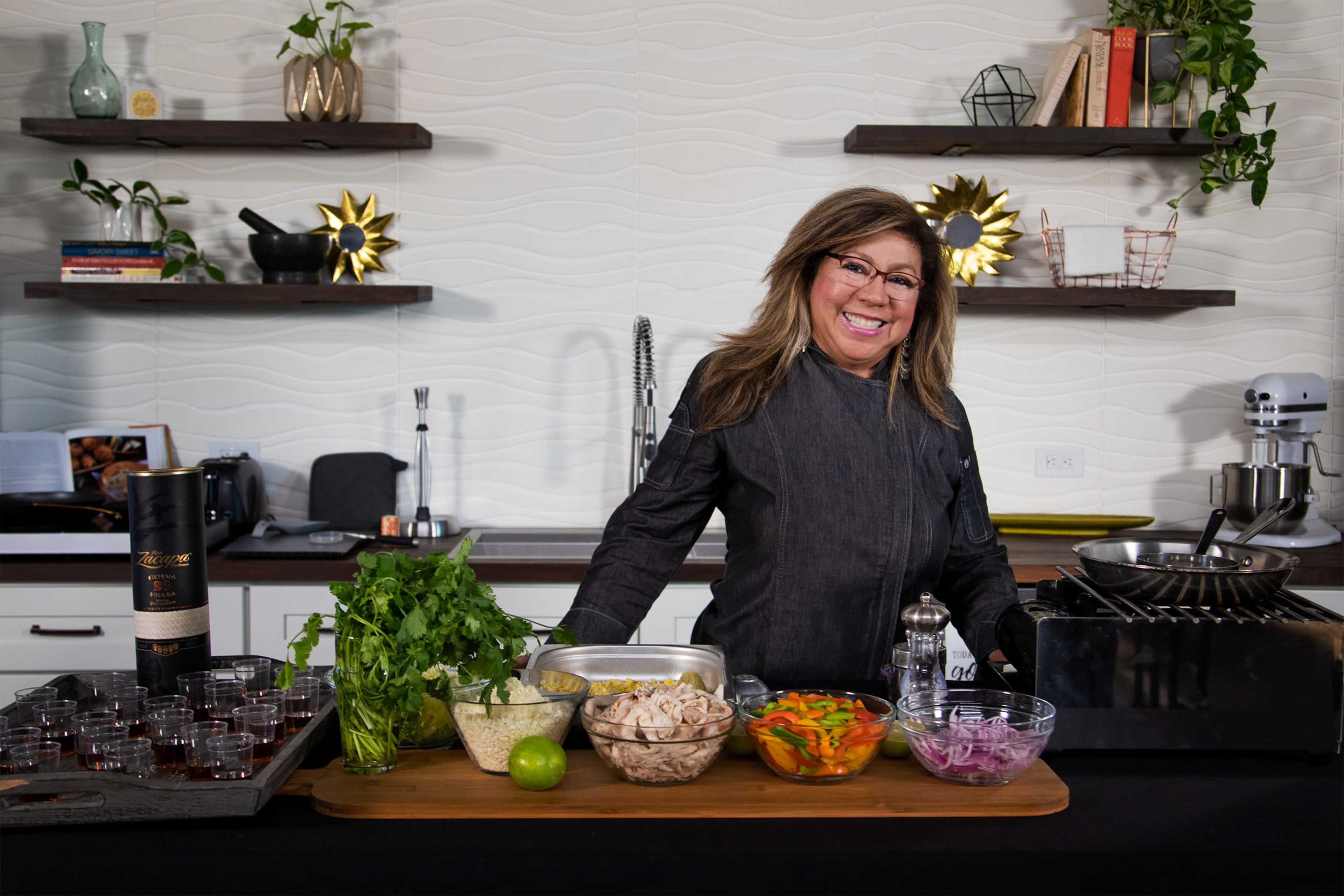 A woman stands behind a wooden table featuring various ingredients and cooking equipment in a modern kitchen setting.