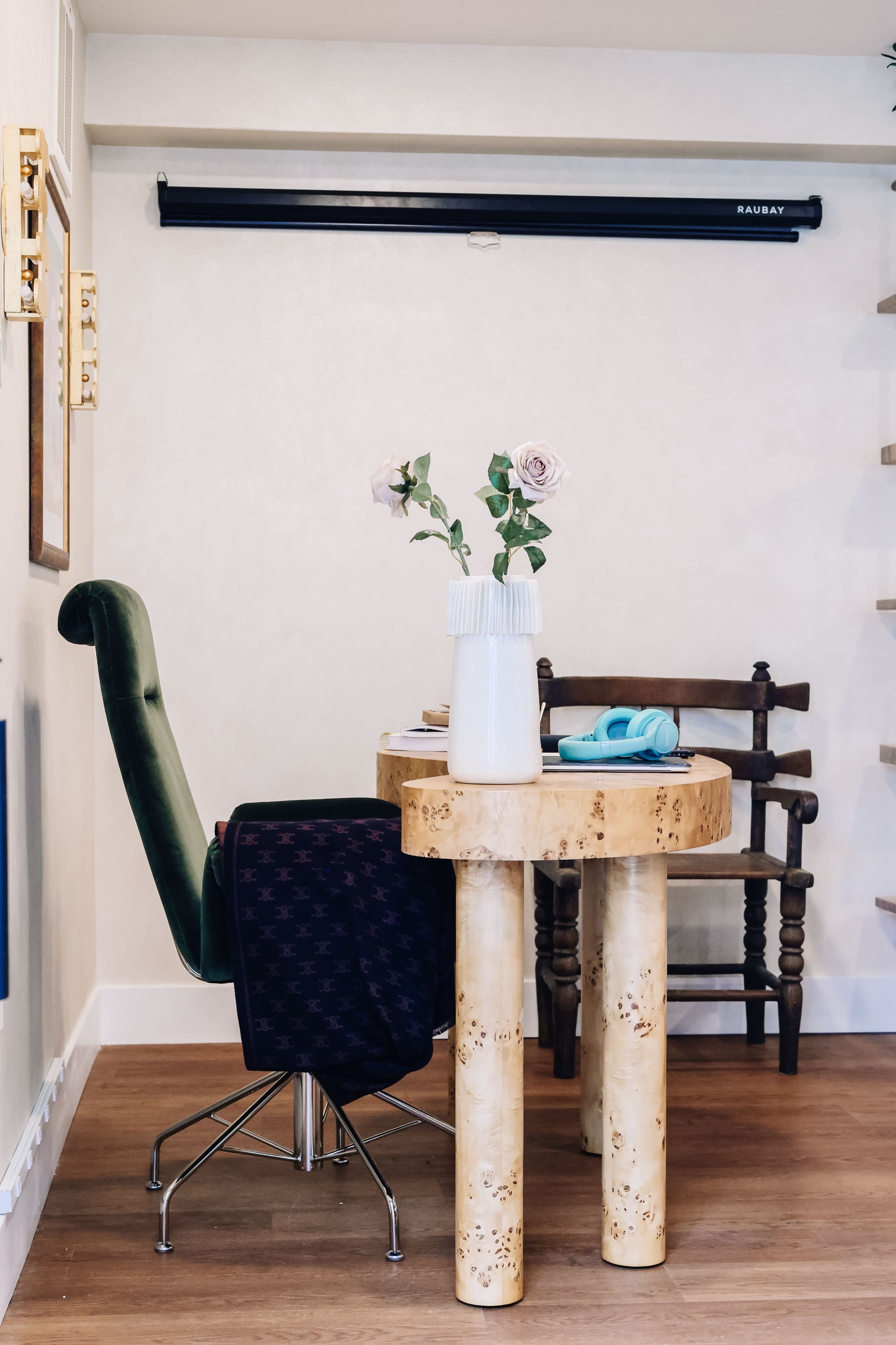 A round wooden table with cylindrical legs sits beside a green chair, with a vase of flowers on the table and a wooden chair in the background.