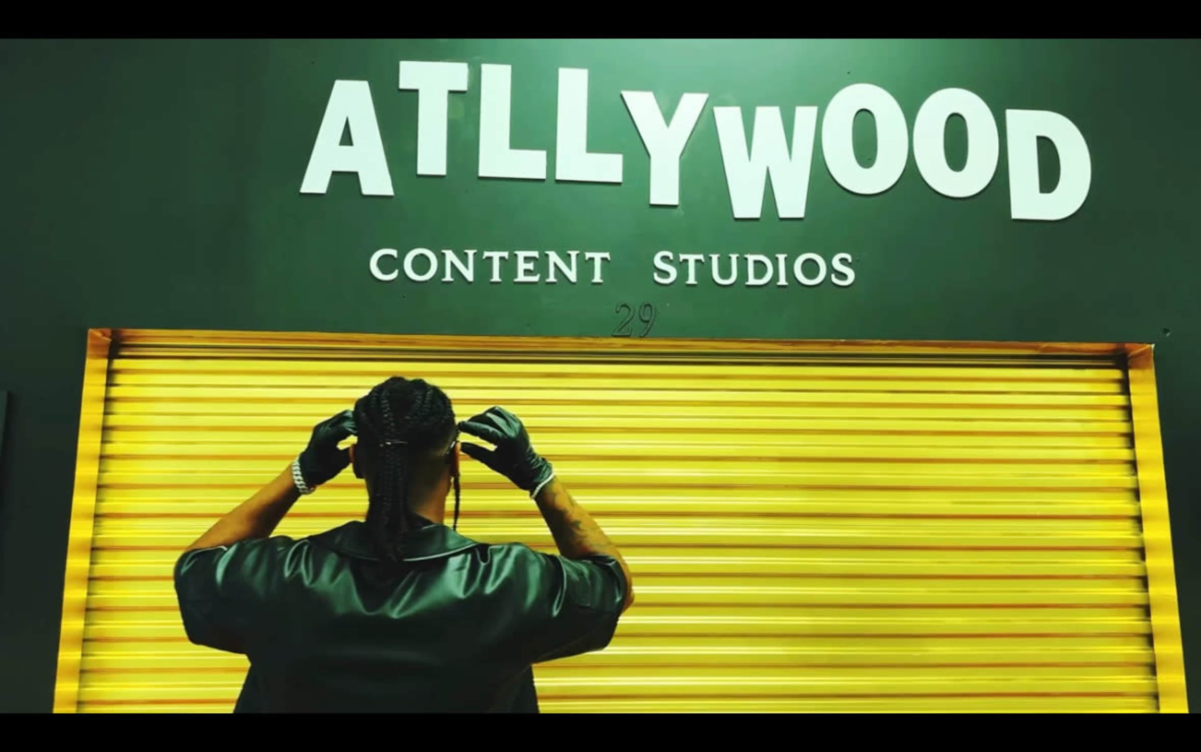 A person with braided hair adjusts their gloves in front of a shutter painted with the words "ATLLYWOOD CONTENT STUDIOS."