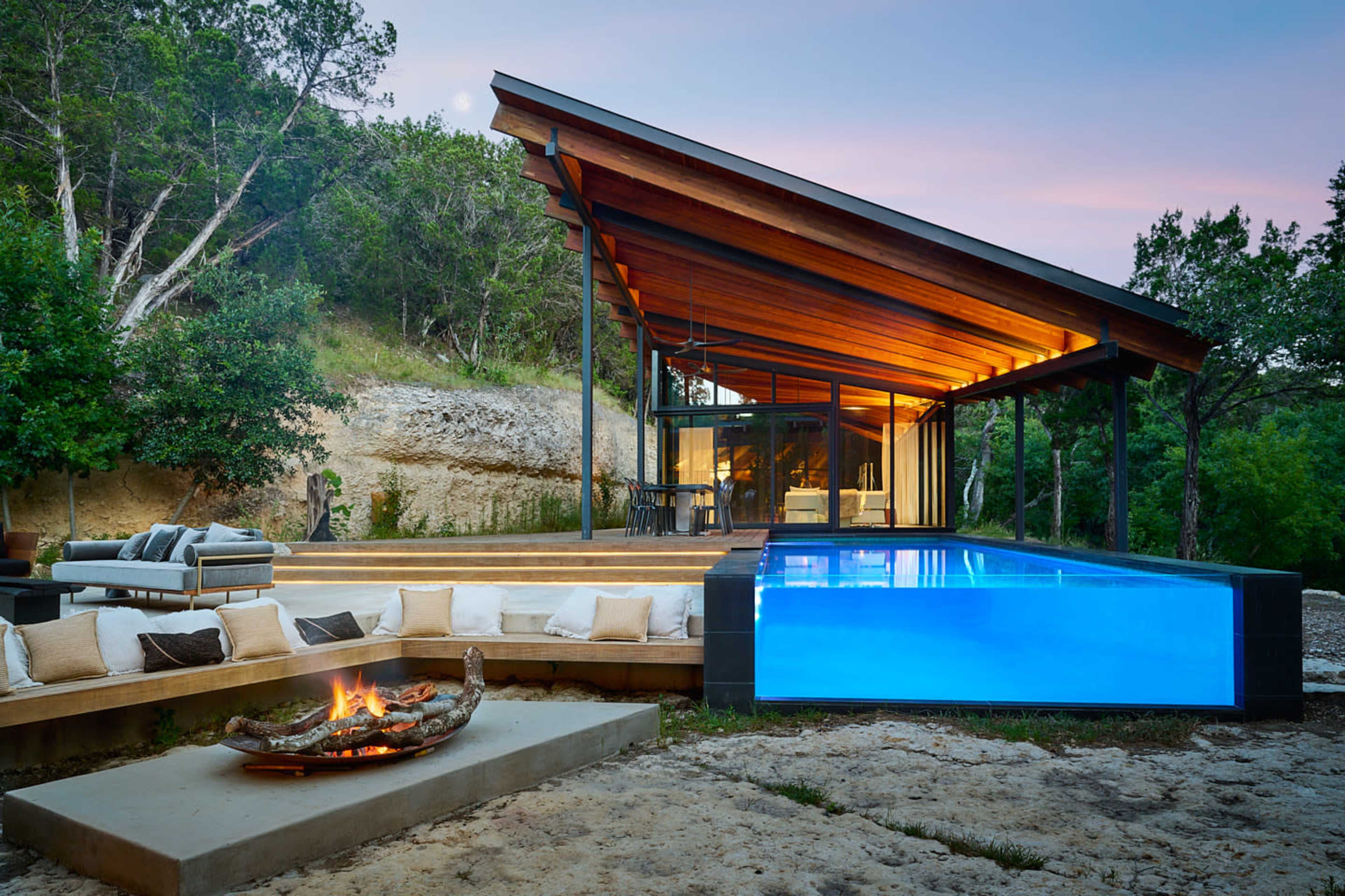 A modern house with a pool and an outdoor seating area is surrounded by trees and rocky terrain.