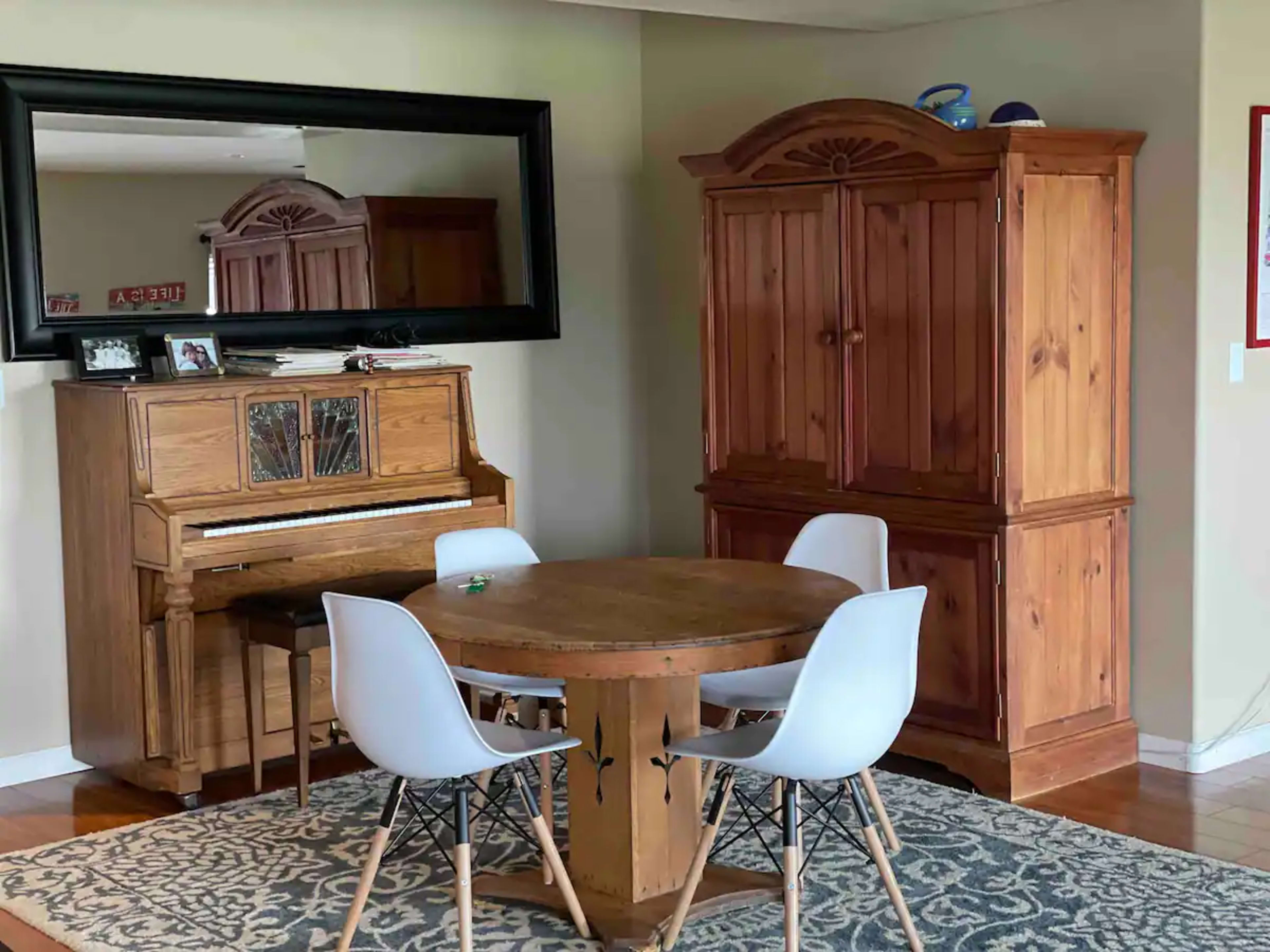 The image shows a dining area with a round table surrounded by four white chairs, a wooden piano to the left, and a wooden cabinet to the right, all set against a wall with a mirror.