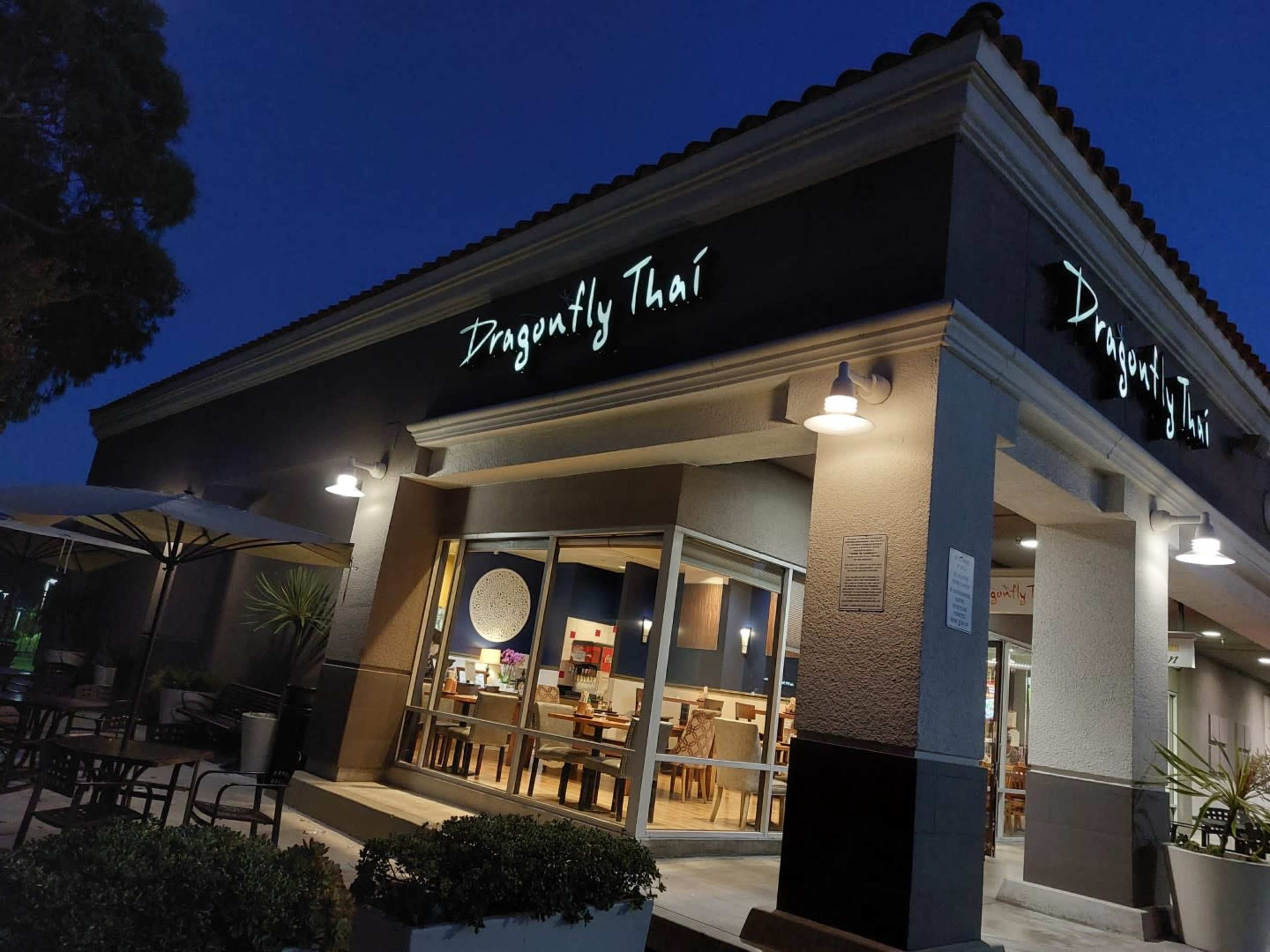 The image shows the exterior of a Thai restaurant called "Dragonfly Thai," featuring a well-lit entrance and outdoor seating under an awning at dusk.