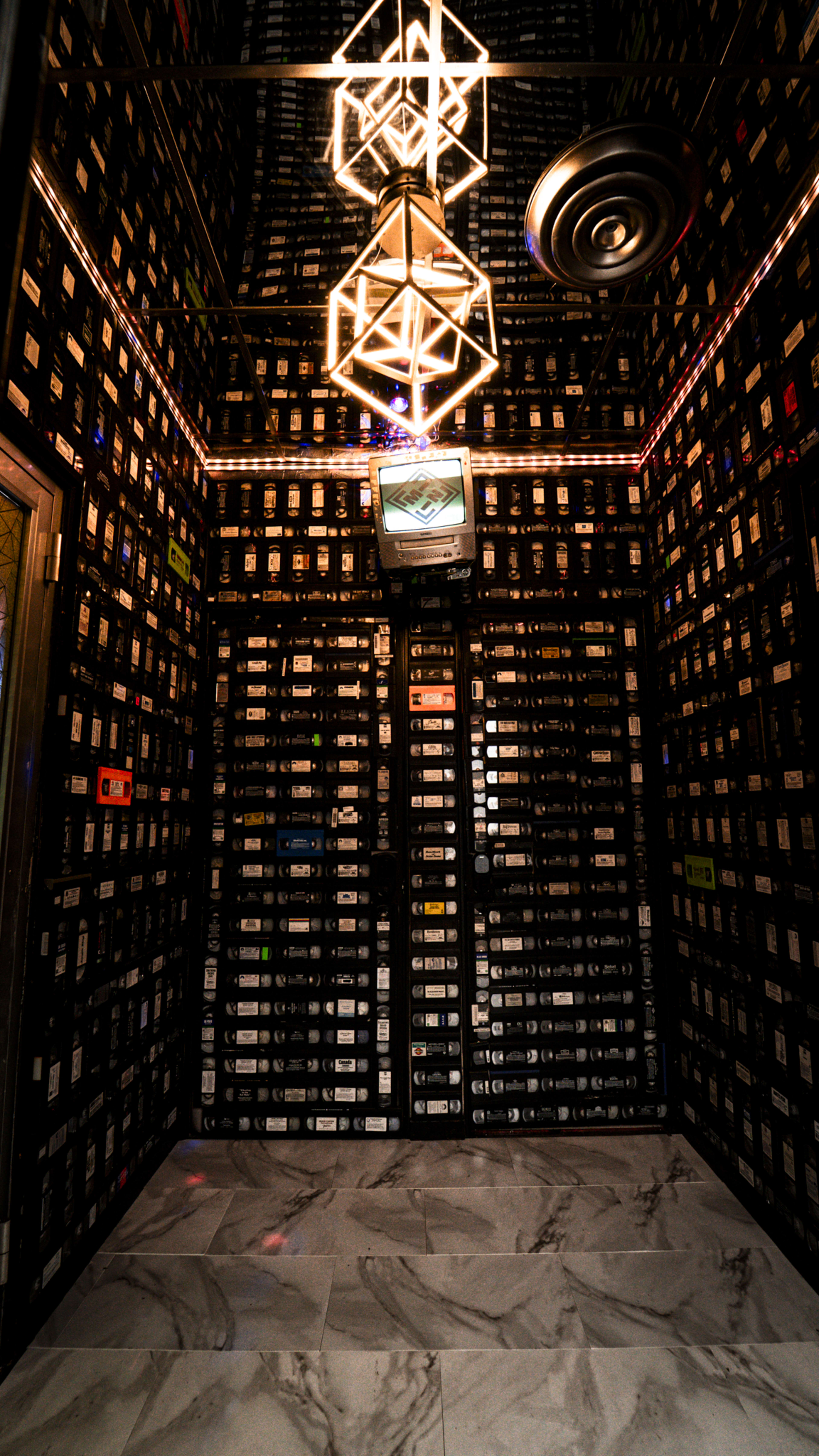 The image shows a dimly lit room with walls covered in cassette tapes and a vintage television mounted in the corner.