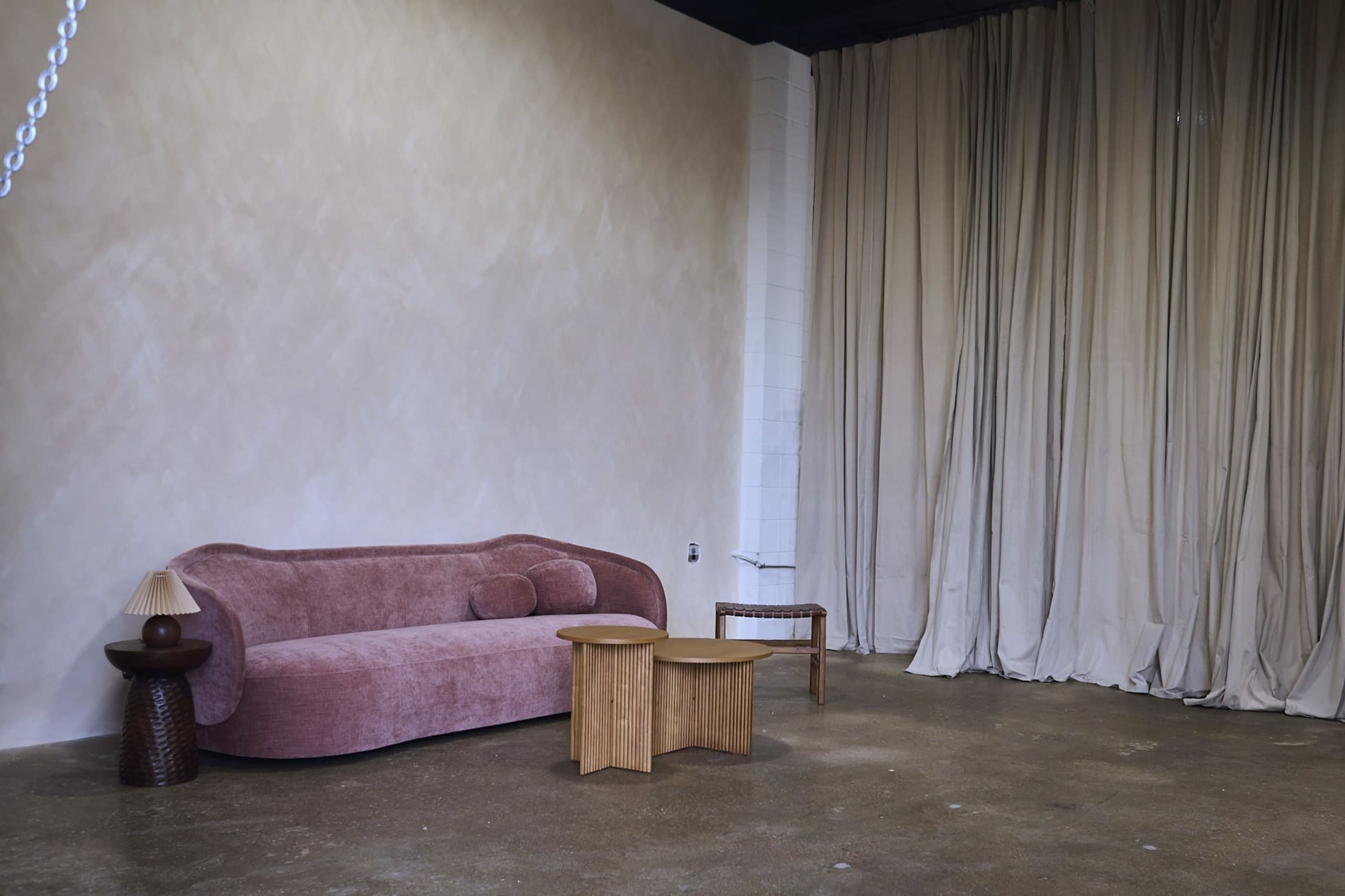 The image features a minimalist interior with a pink sofa, two wooden side tables, and a curtain backdrop.