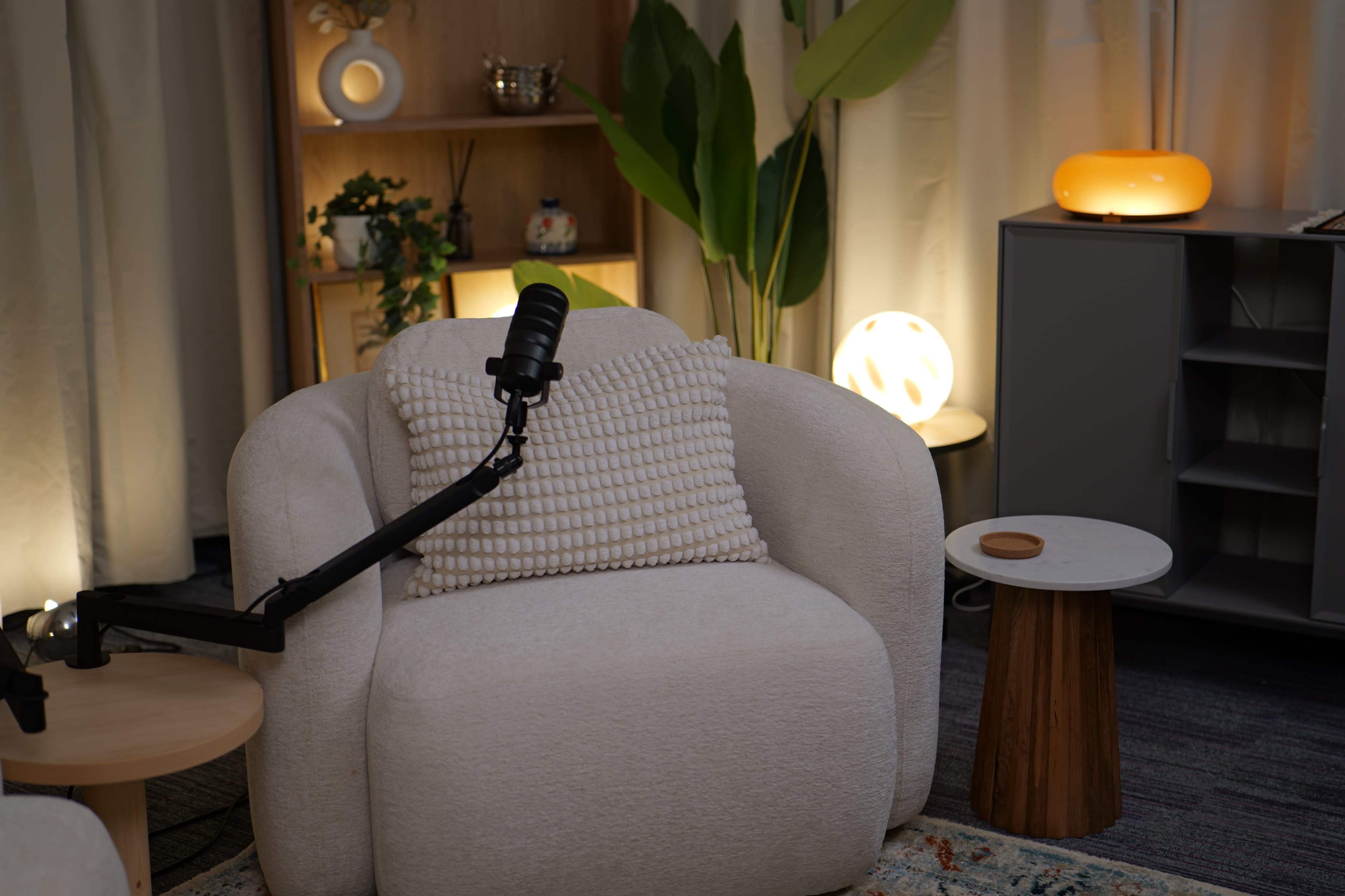 A soft, white armchair with a microphone attached to an arm stands beside a small wooden table and various decorative plants and lamps.