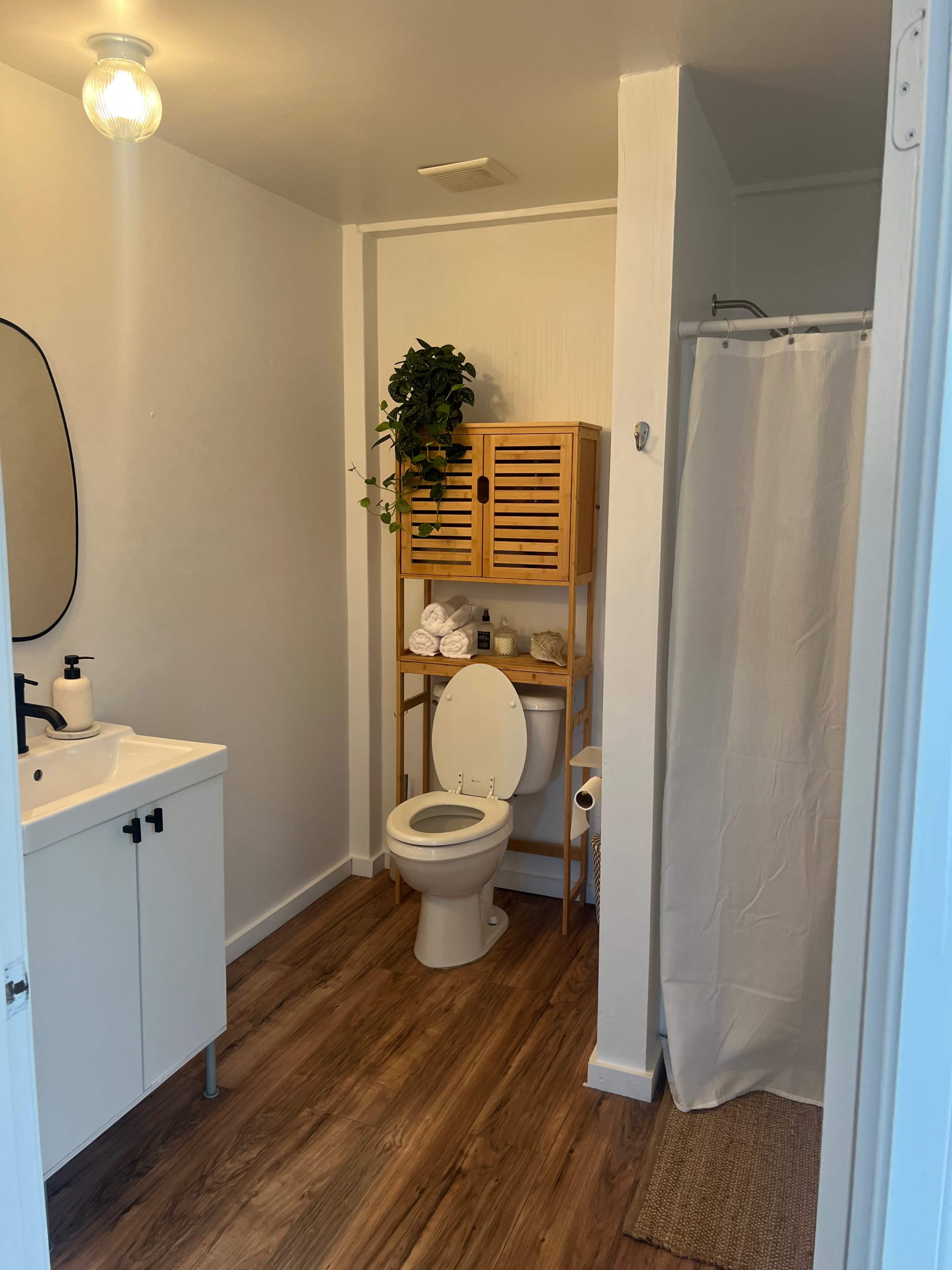 A bathroom with a white sink and cabinet, a wooden shelving unit with towels and plants, a toilet, and a shower curtain.