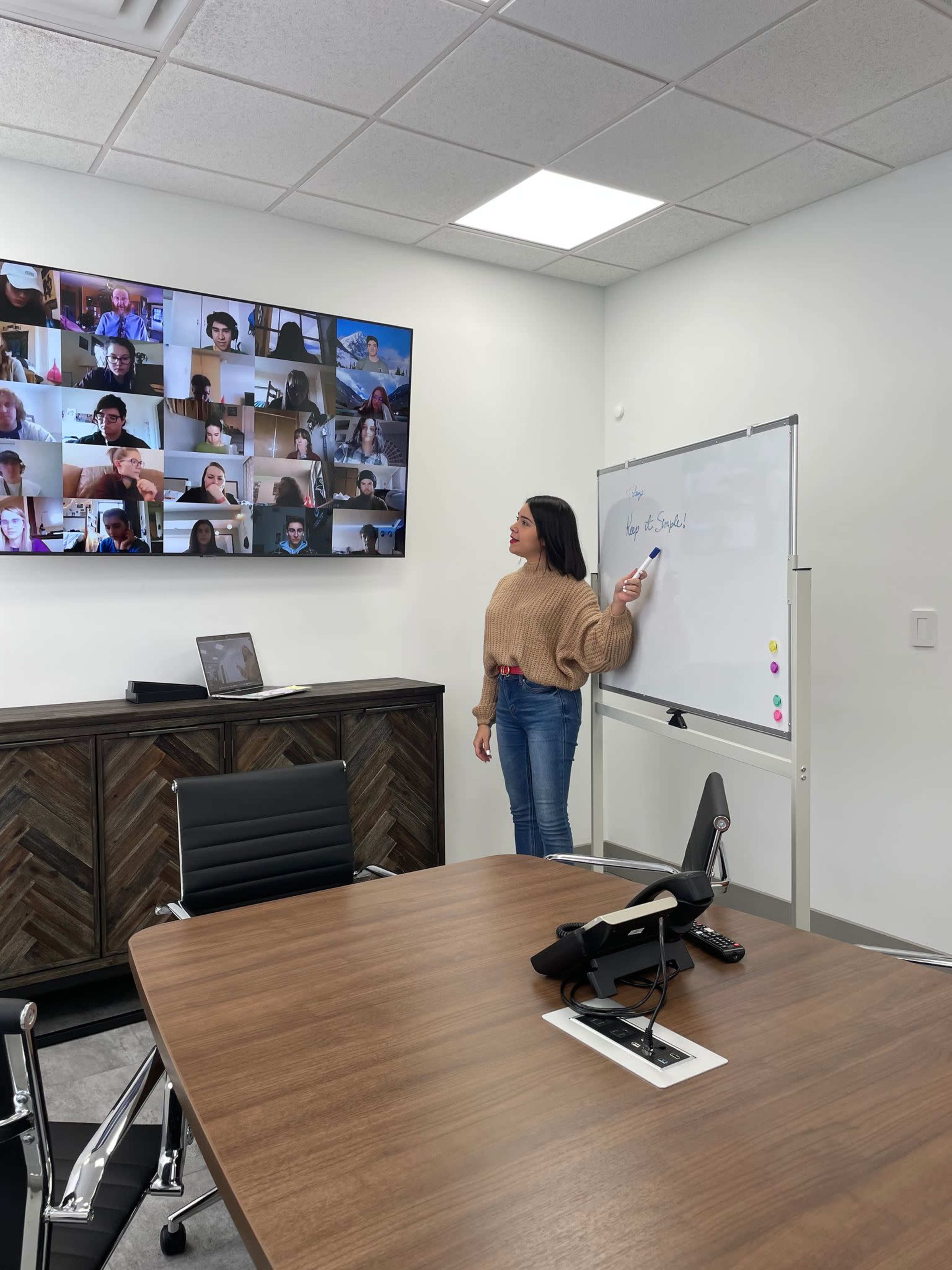 A person stands by a whiteboard in a conference room while a video call with multiple participants is displayed on a screen behind them.