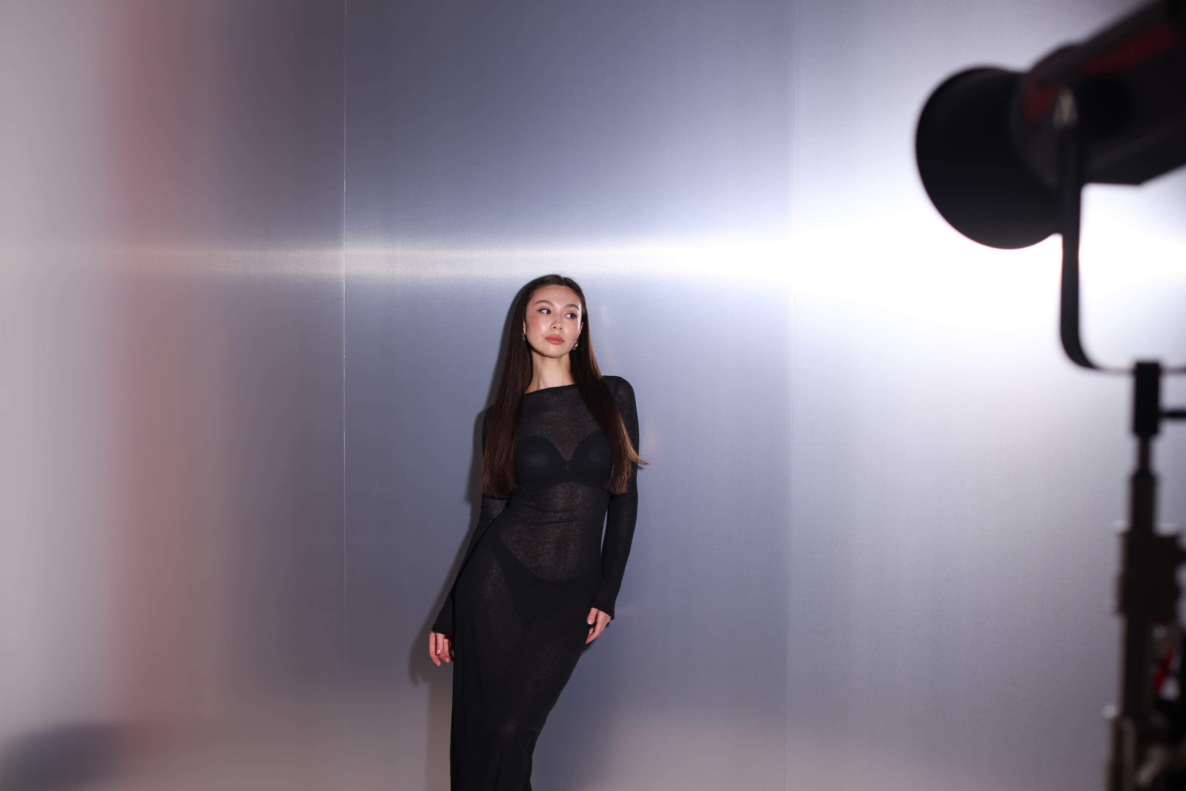 A woman poses in a black outfit against a metallic backdrop, with studio lighting illuminating the scene.