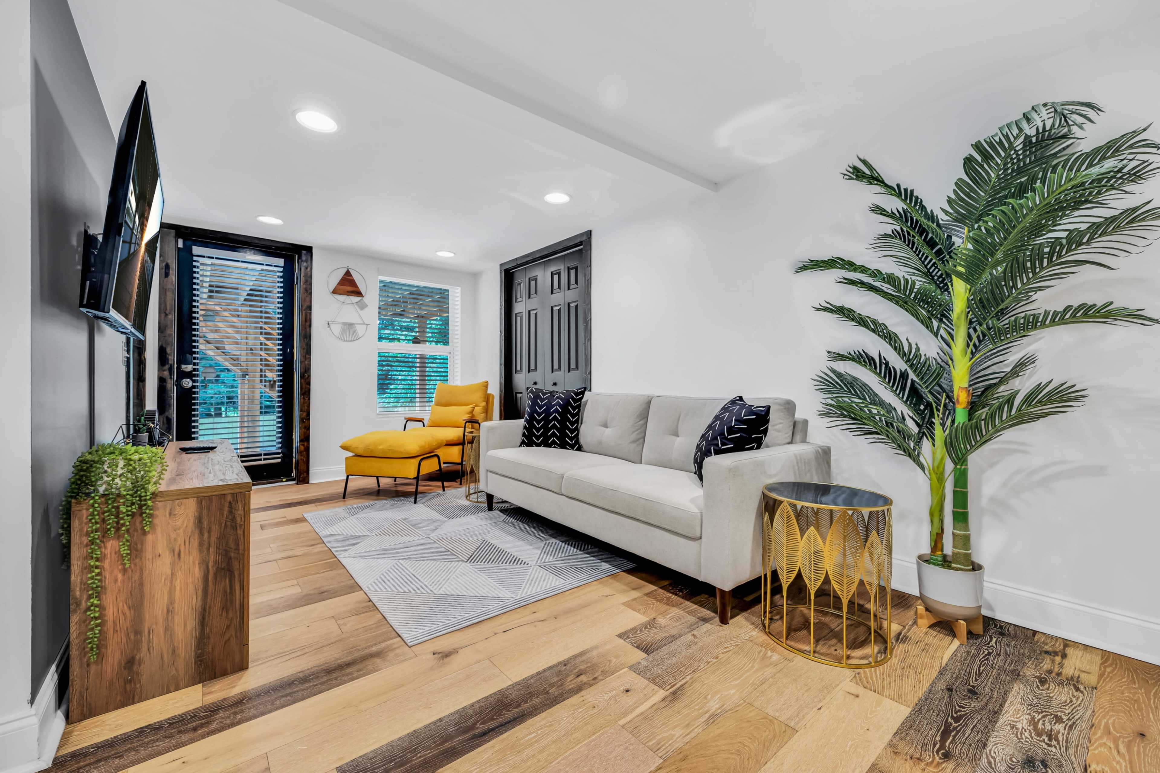 A modern living room with a gray sofa, a yellow armchair, a wooden TV stand, and large windows with blinds, featuring a potted plant in one corner.