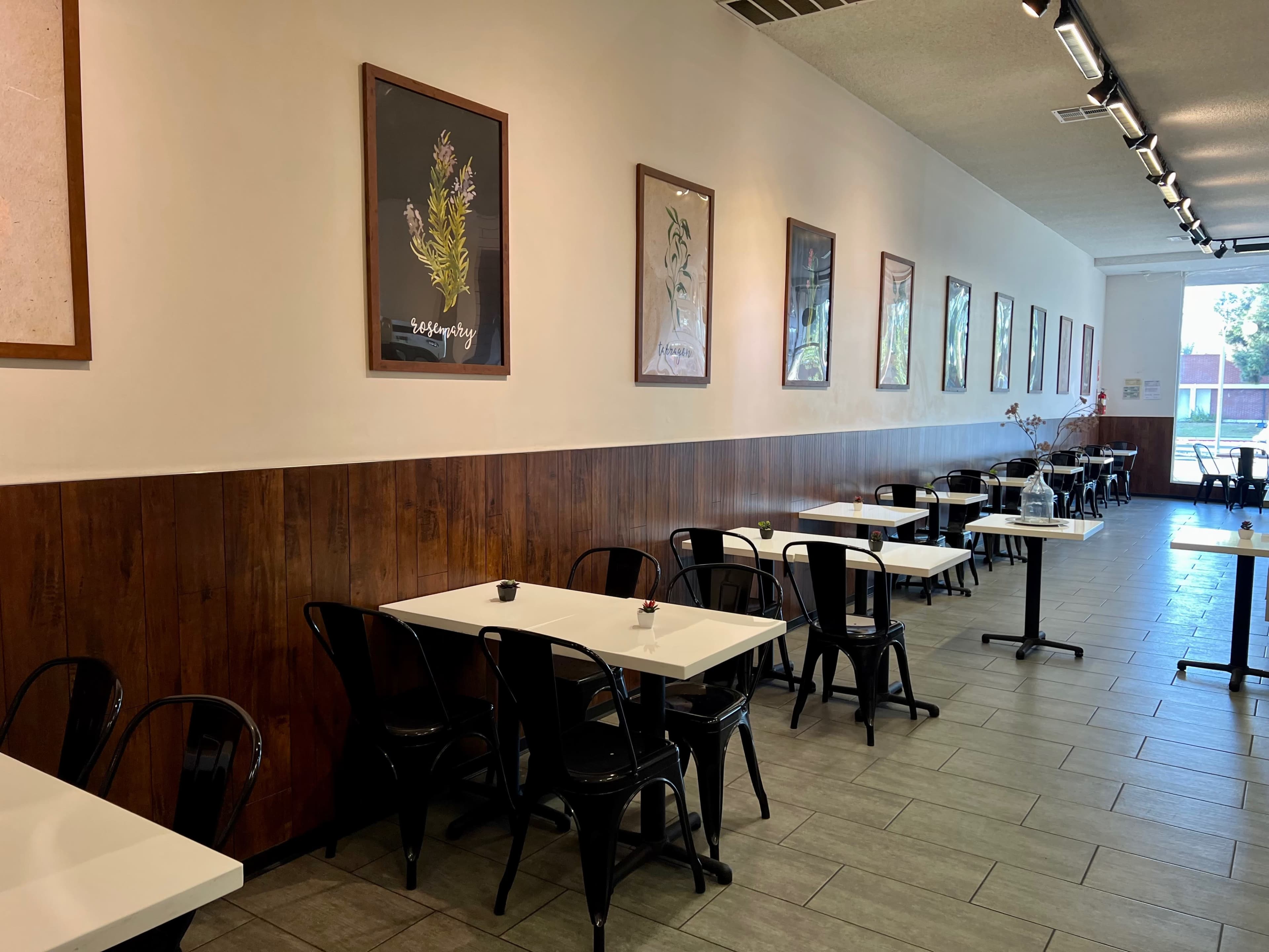 The image shows a modern café interior featuring white tables and black chairs along a wooden-paneled wall adorned with framed artwork.