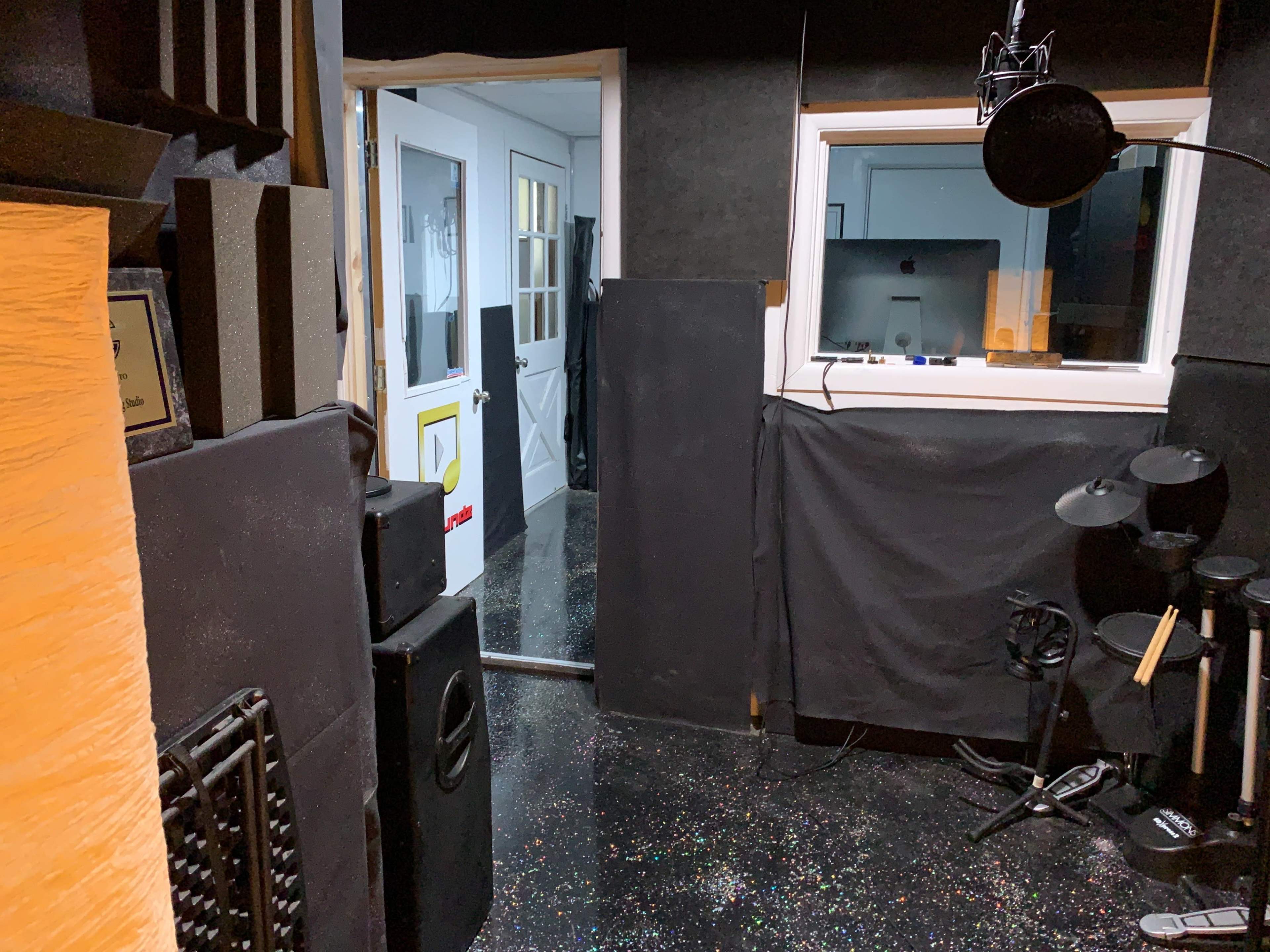 The image shows a recording studio with soundproofing foam on the walls, a microphone setup, and a door leading to a separate room.