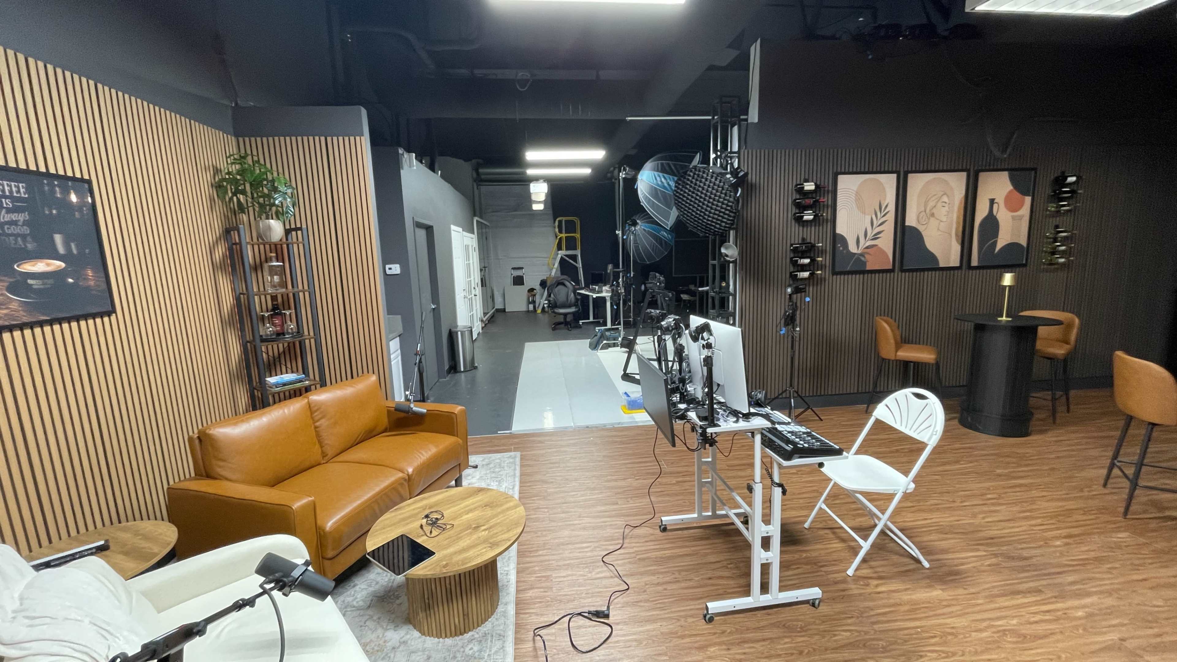 The image shows a modern studio space featuring a brown leather couch, a round wooden coffee table, a white folding chair, and a set-up for video production with cameras and lighting equipment.