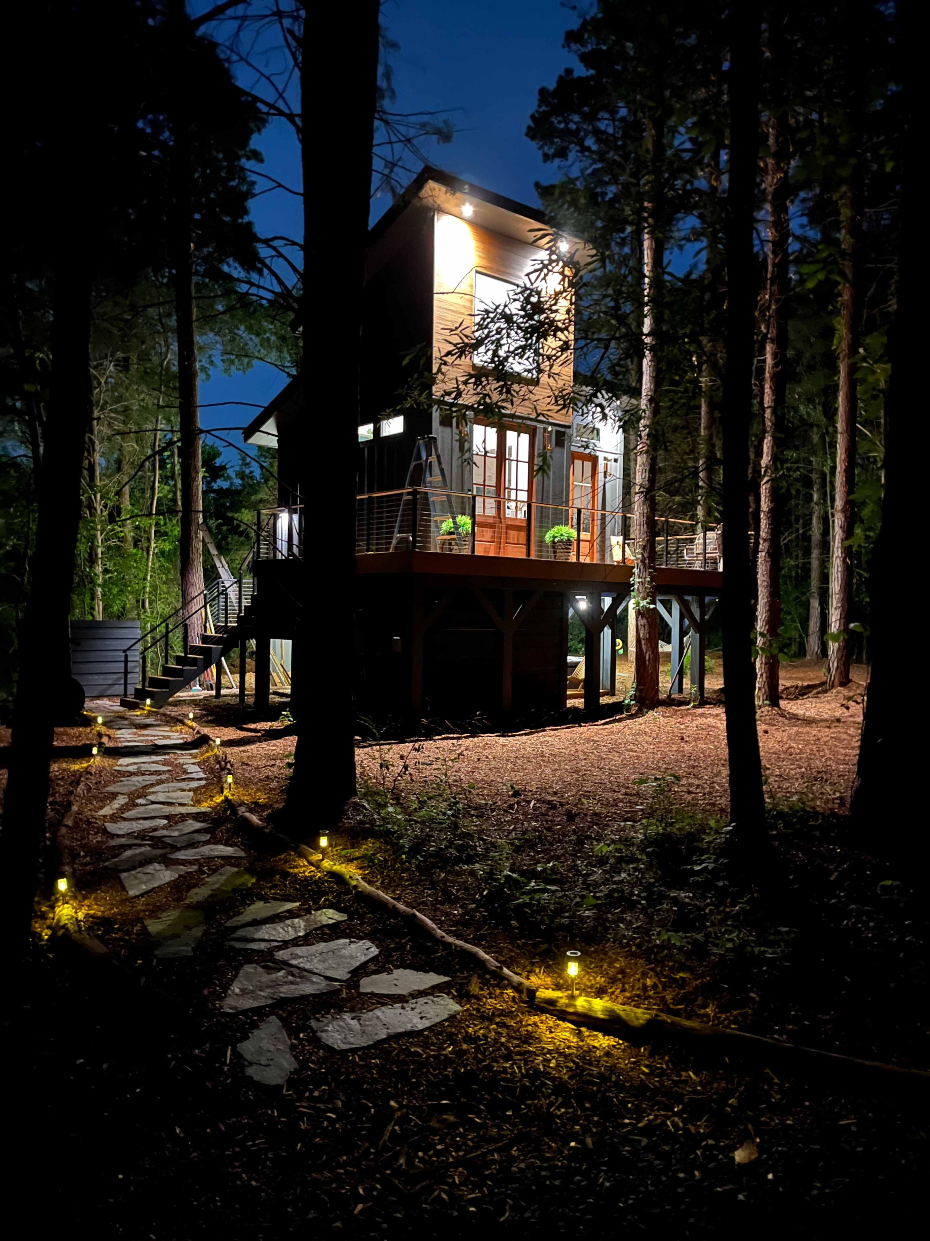 A modern two-story cabin is illuminated at night, surrounded by tall trees and a stone pathway lined with solar lights.