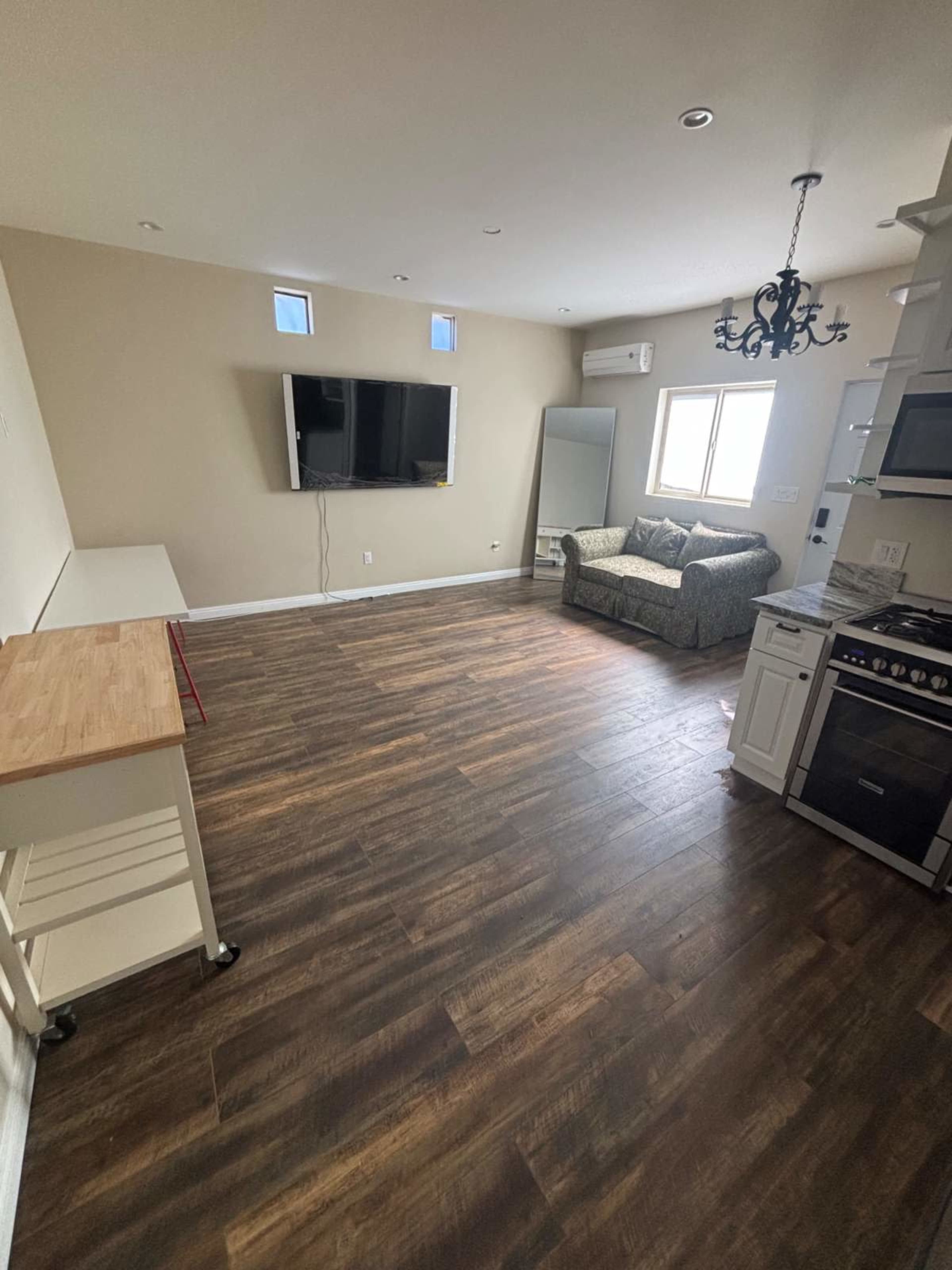 The image shows a living area with hardwood floors, a sofa, a TV mounted on the wall, and a kitchen area visible in the corner.