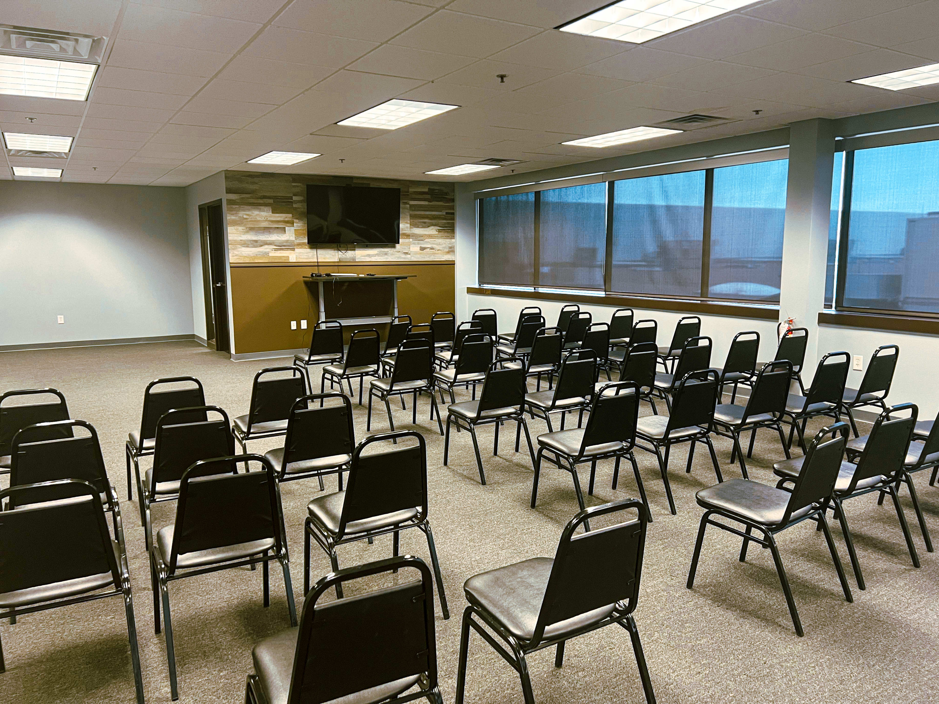 The image shows a meeting room arranged with rows of black chairs facing a television screen mounted on the wall.