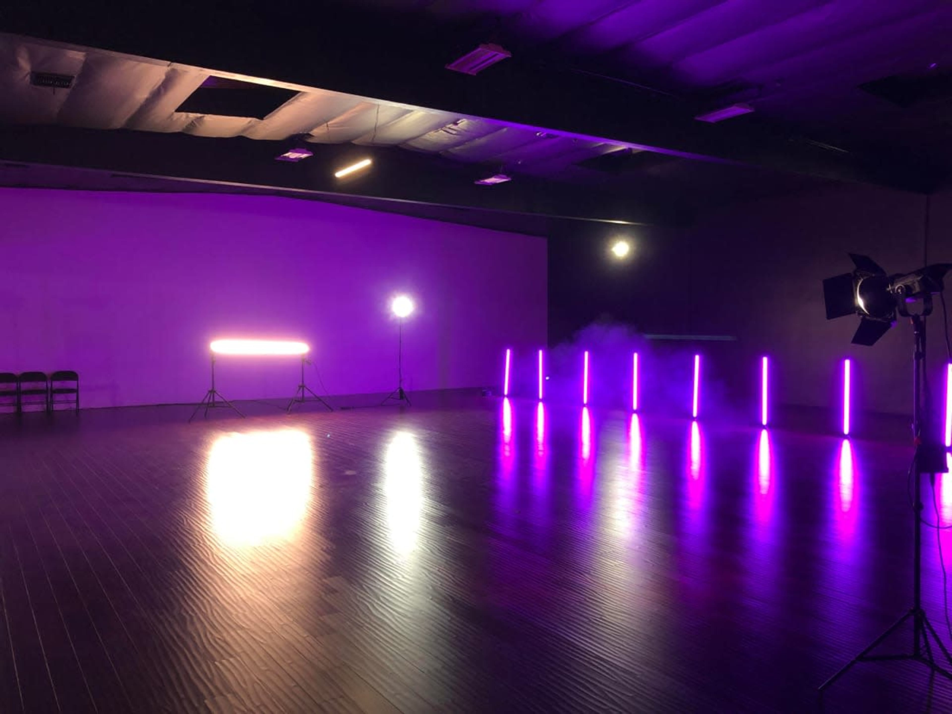An empty room with purple lighting, featuring two light sources and a row of illuminated poles against the back wall.