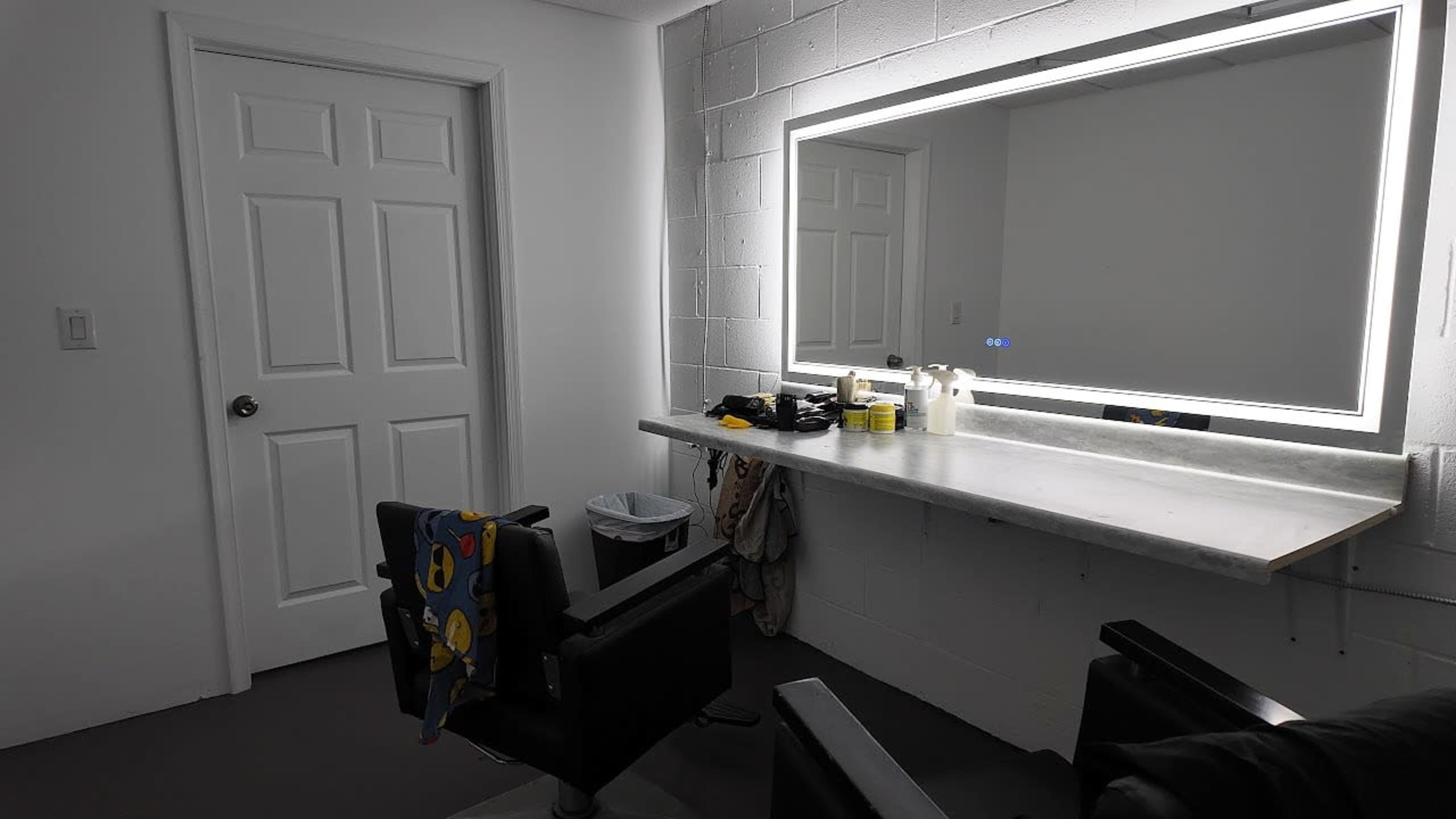 The image shows a hair salon area with a countertop, a backlit mirror, and two black salon chairs in front of it, along with a door and a trash can in the background.