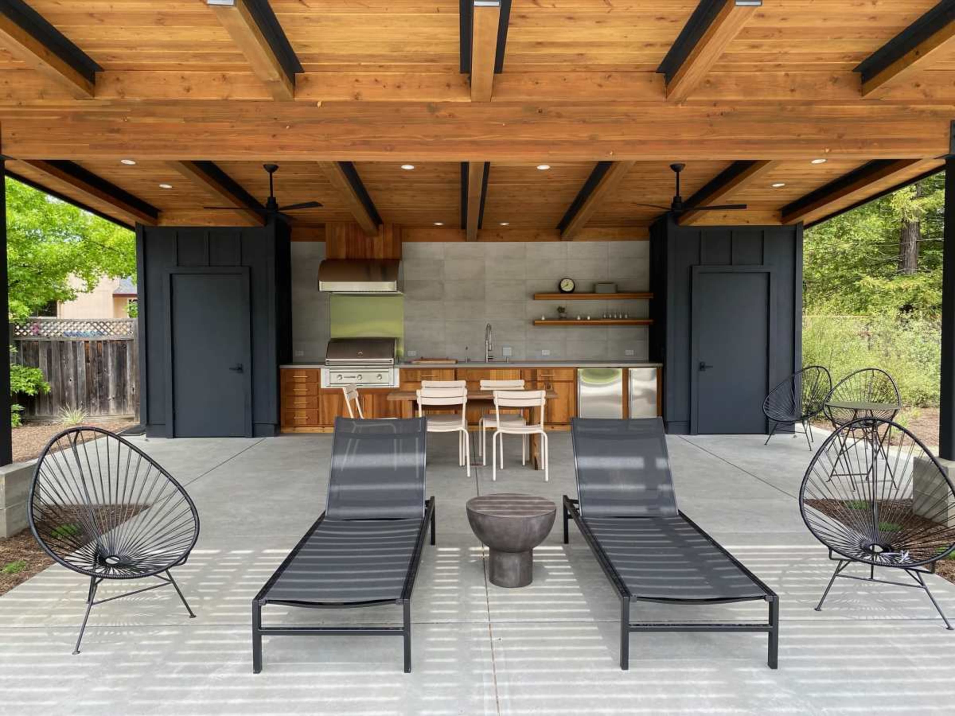 The image shows a covered outdoor space with two lounge chairs, a dining table, and a kitchen area featuring stainless steel appliances.