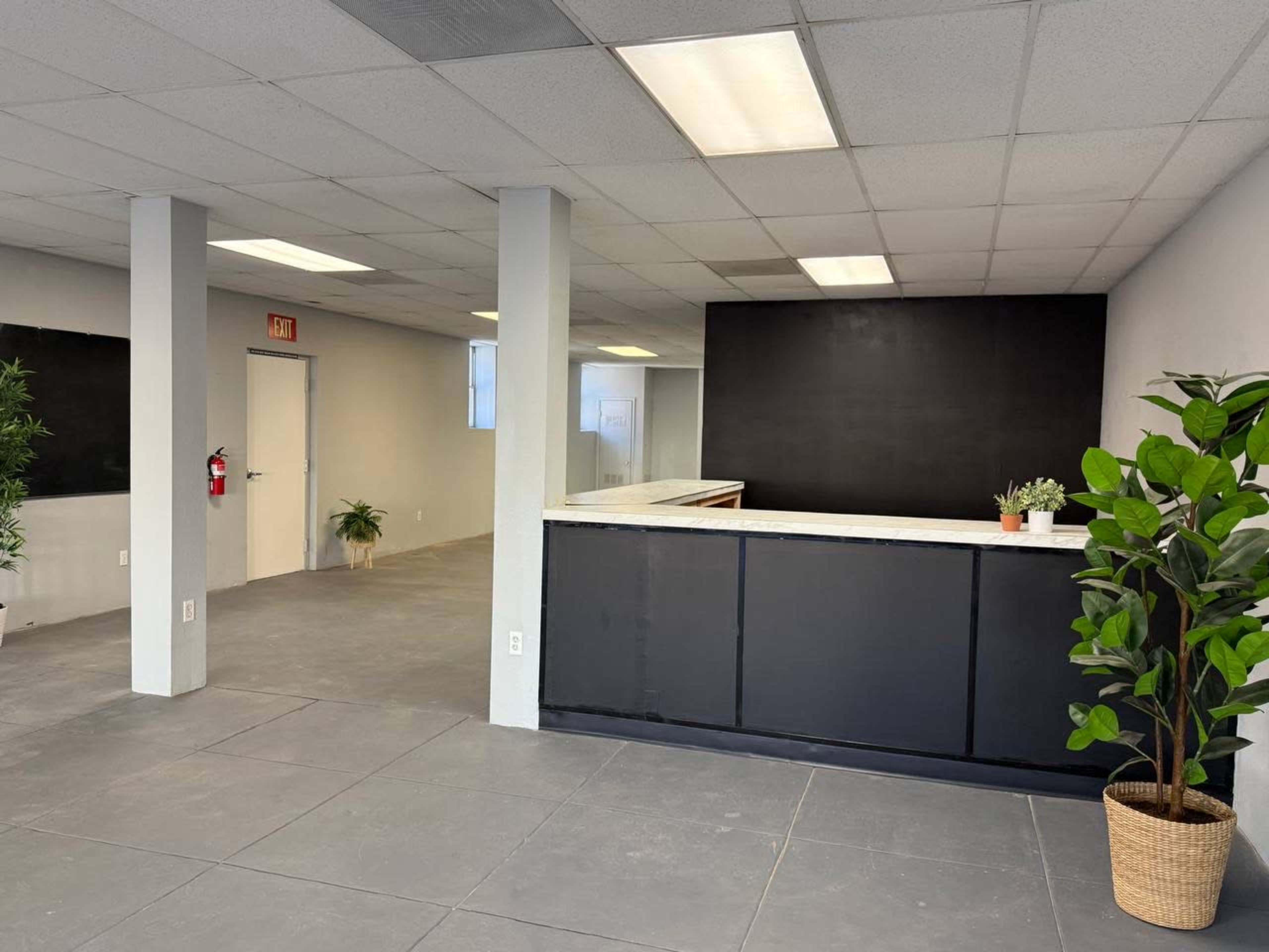 The image shows a spacious, empty office interior with a reception desk, gray tile flooring, and potted plants in the corners.