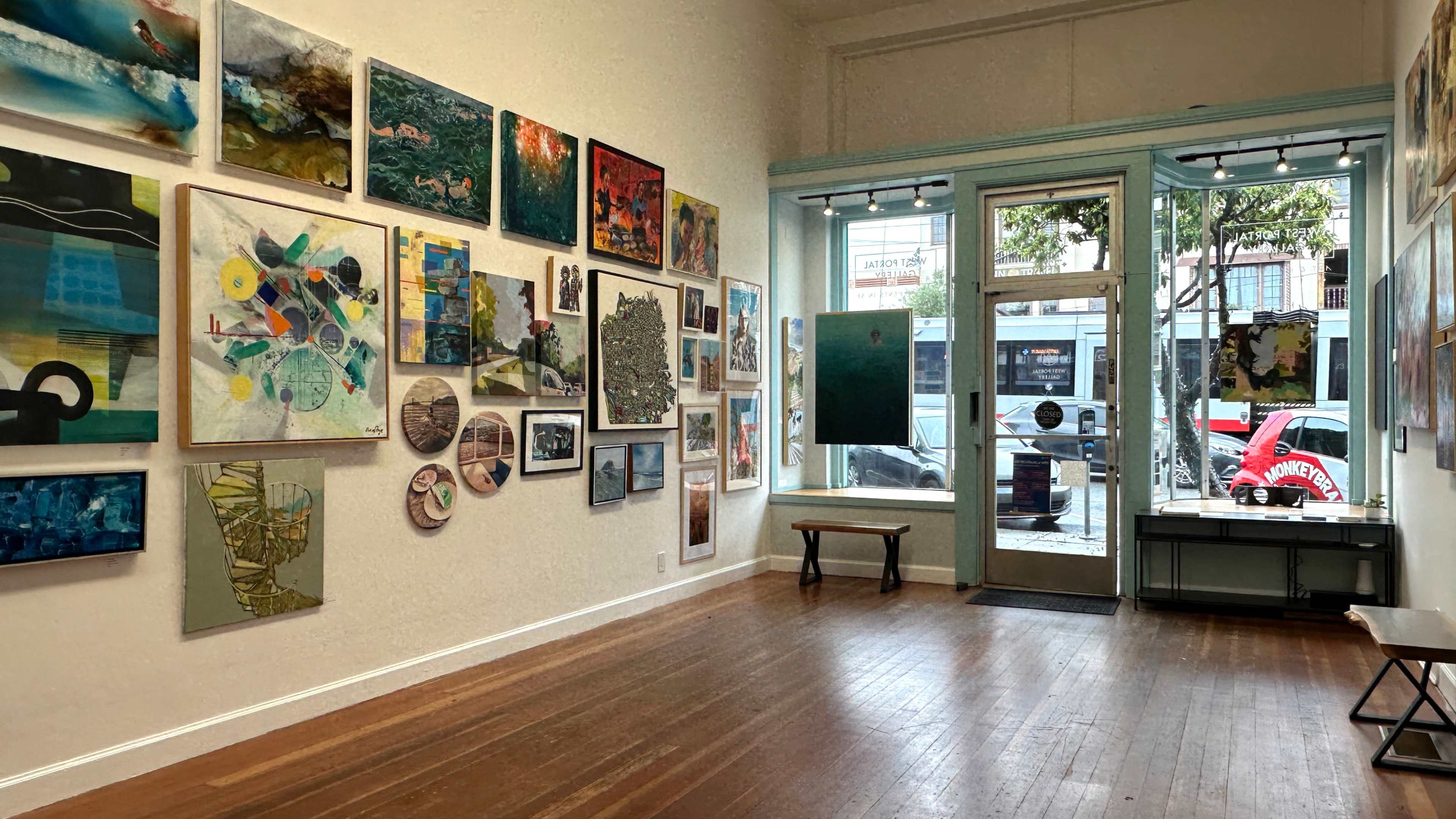 The image shows an art gallery with various artworks displayed on the walls and a bench in the center of the wooden floor.