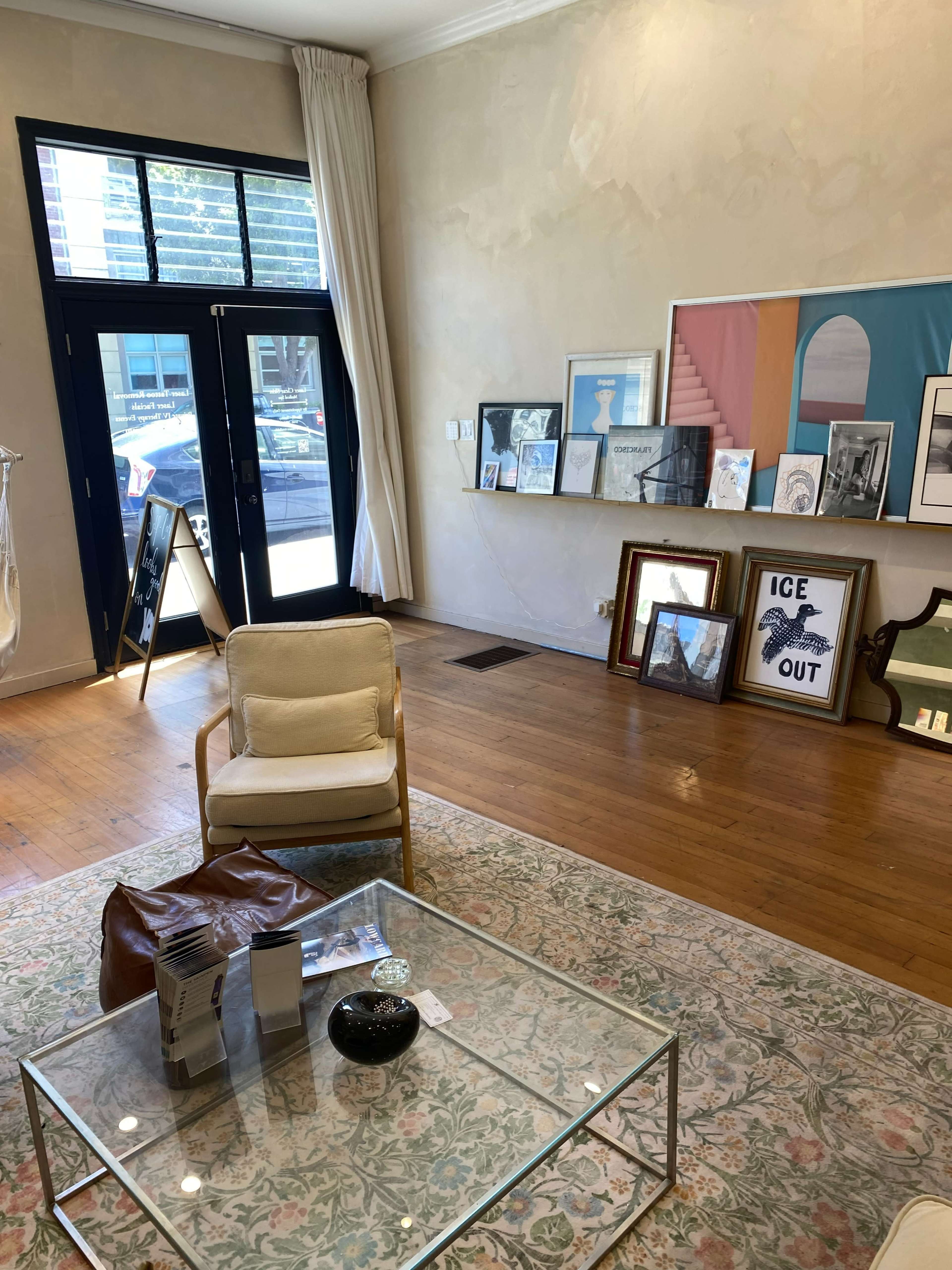 The image shows a room with wooden flooring, a glass coffee table, an armchair, and several framed artworks displayed against the walls.