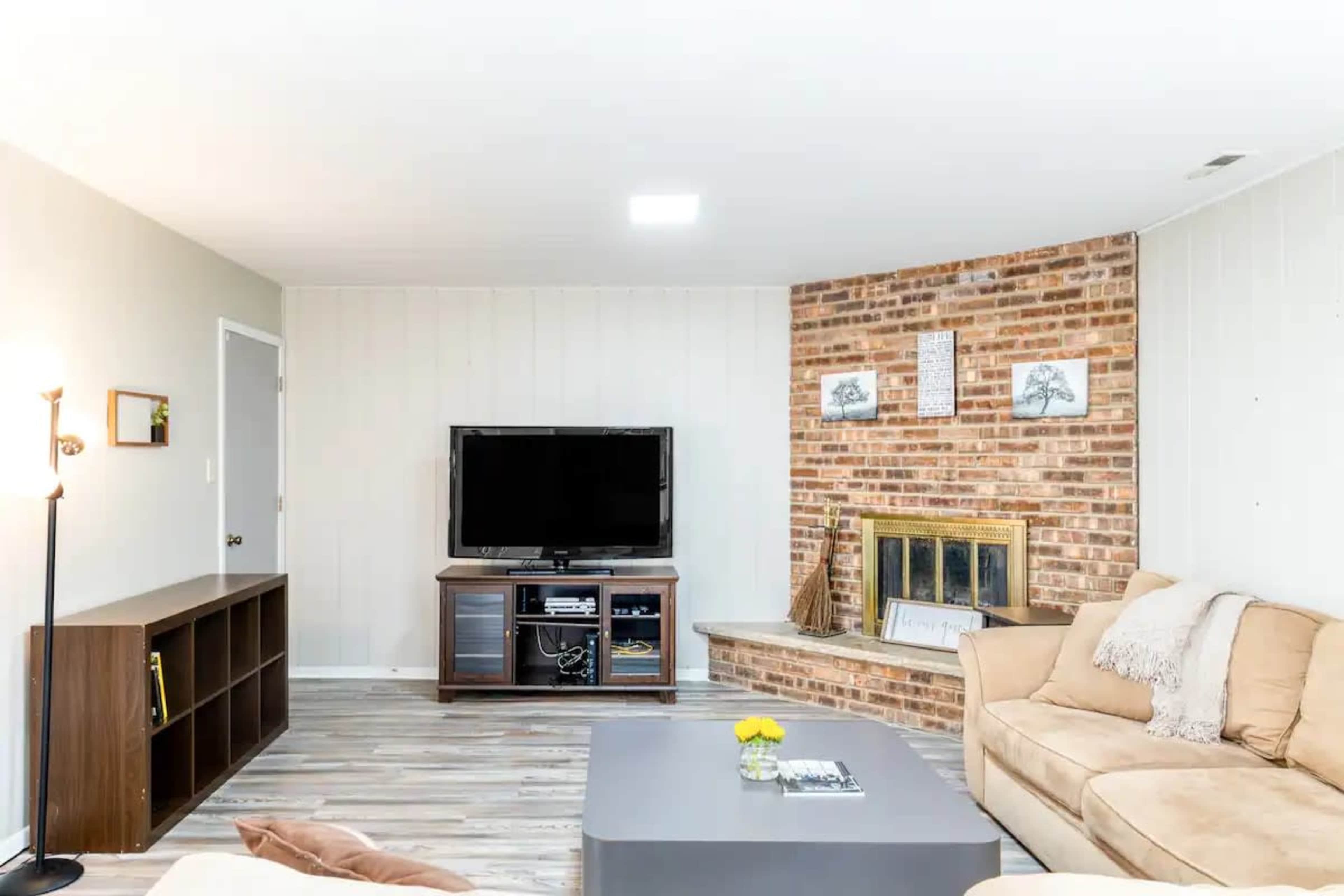 The image shows a modern living room featuring a seating area with a beige sofa, a coffee table, a television mounted on the wall, and a fireplace with a brick surround.