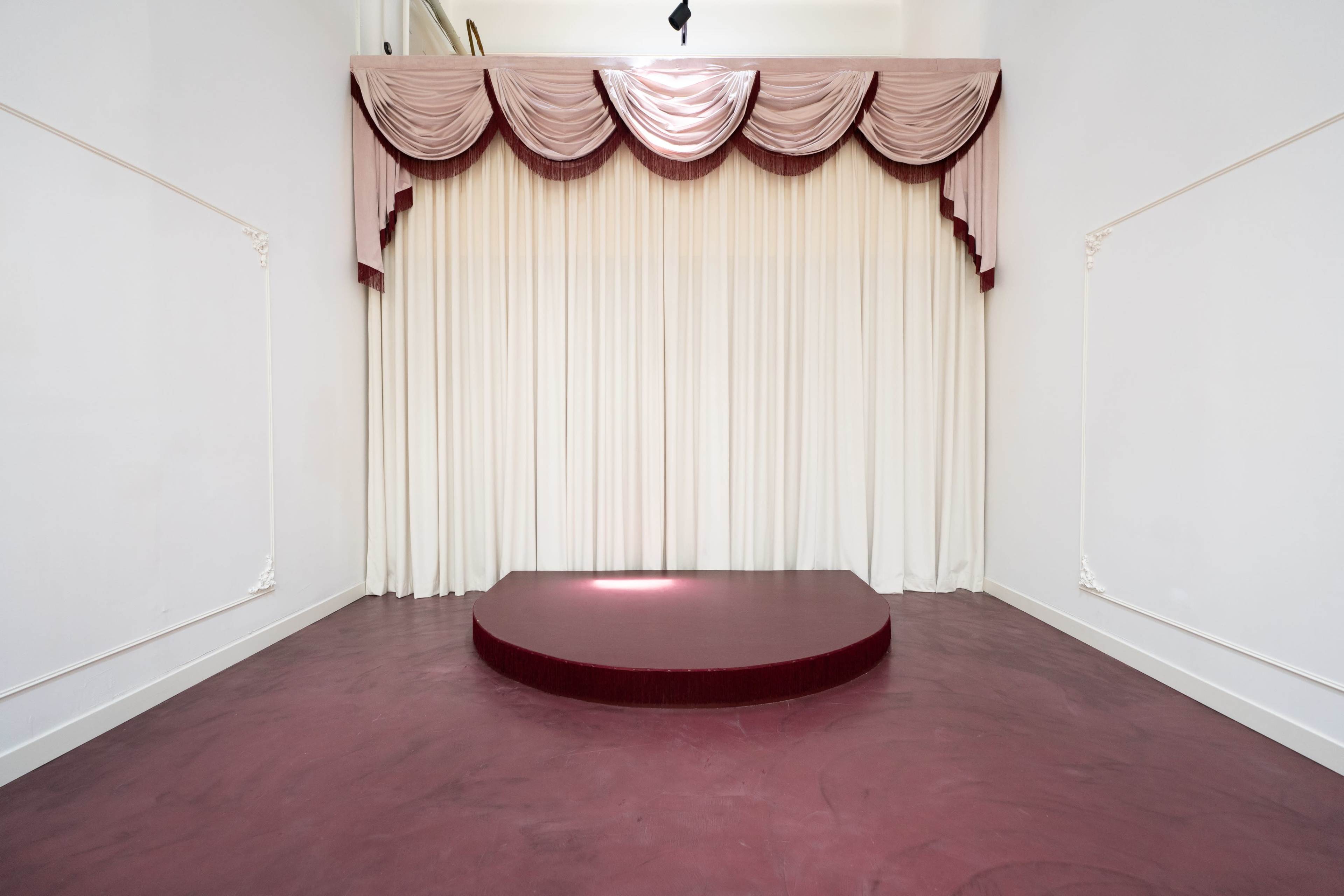 The image shows a small stage with a circular platform in front of a backdrop of white curtains and burgundy drapery.