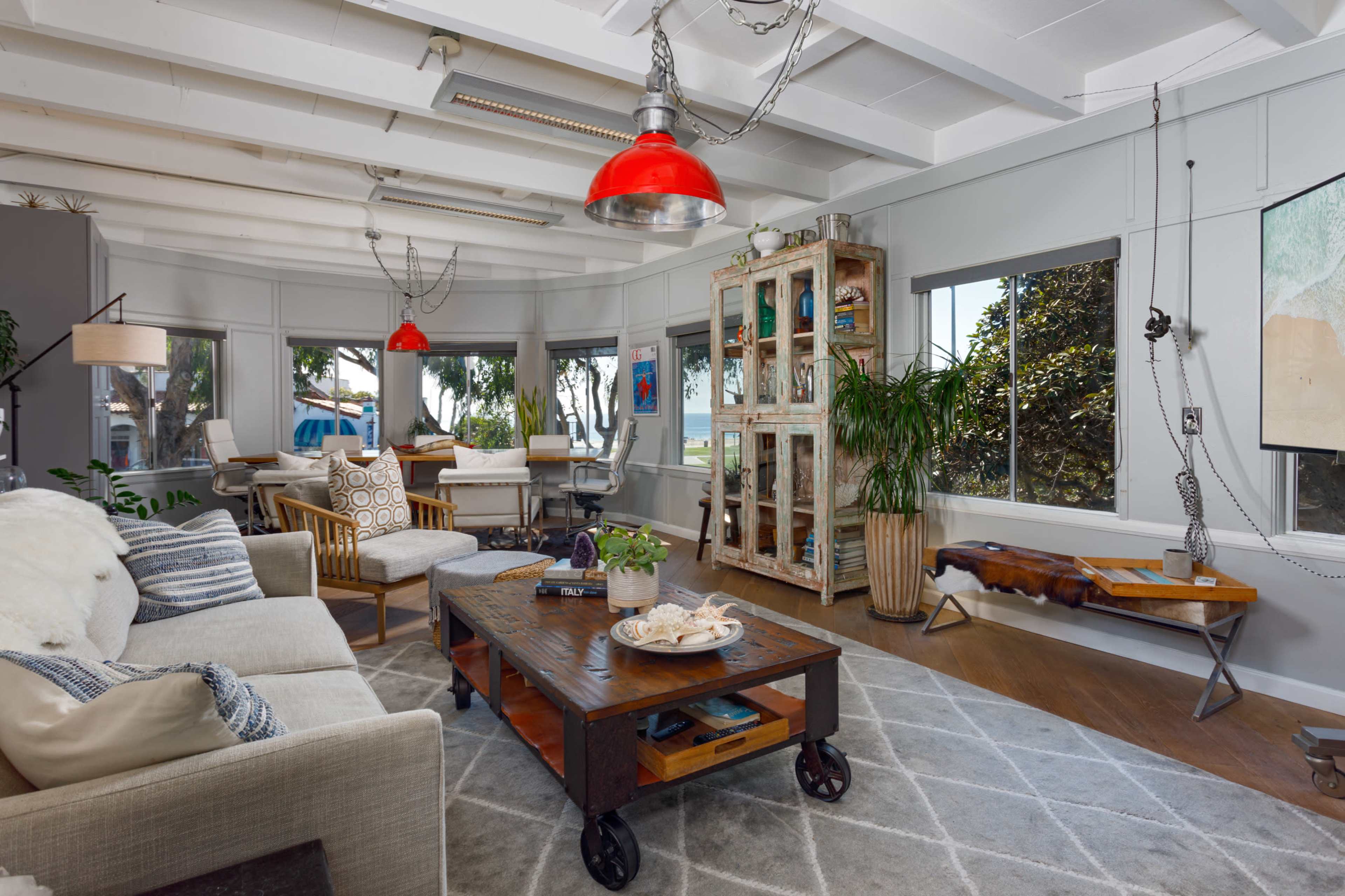The living room features a mix of contemporary furniture, a large coffee table on wheels, and large windows that offer a view of greenery outside.