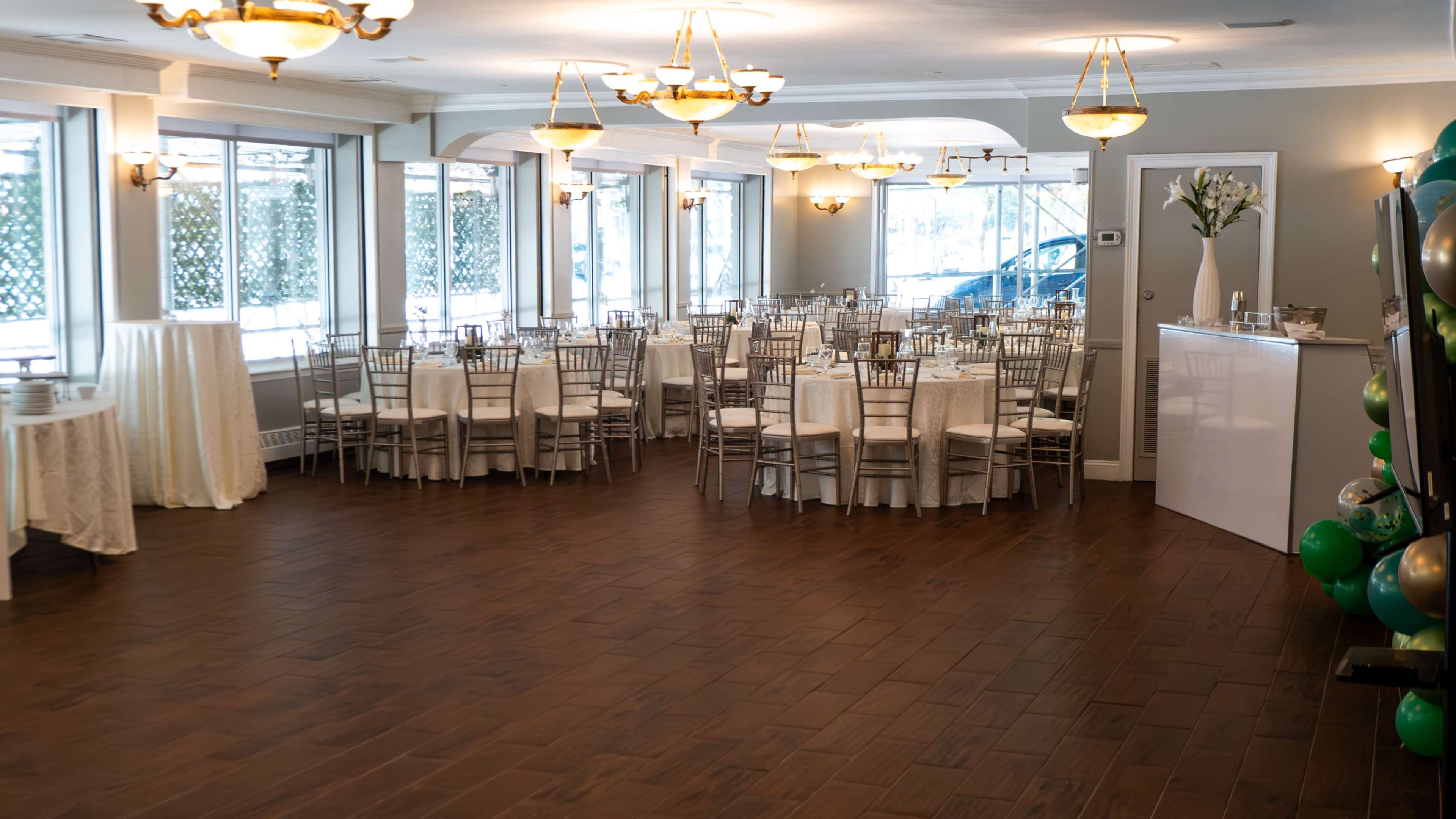 A banquet hall is set up with white-tableclothed tables and chairs arranged for an event, featuring large windows that allow natural light to fill the space.