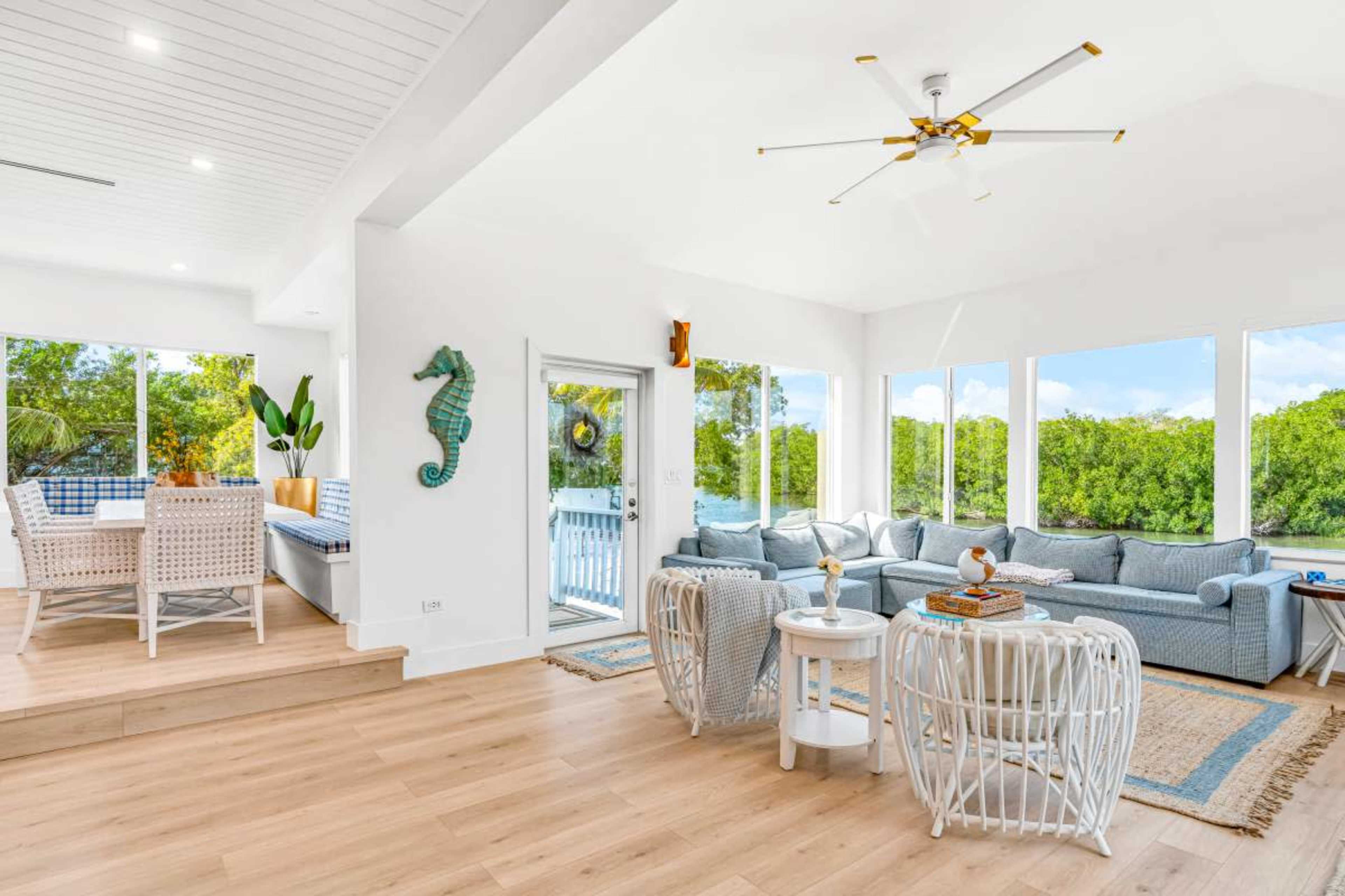 The image shows a bright, modern living room with large windows, light wood flooring, and comfortable furniture, overlooking a natural landscape.