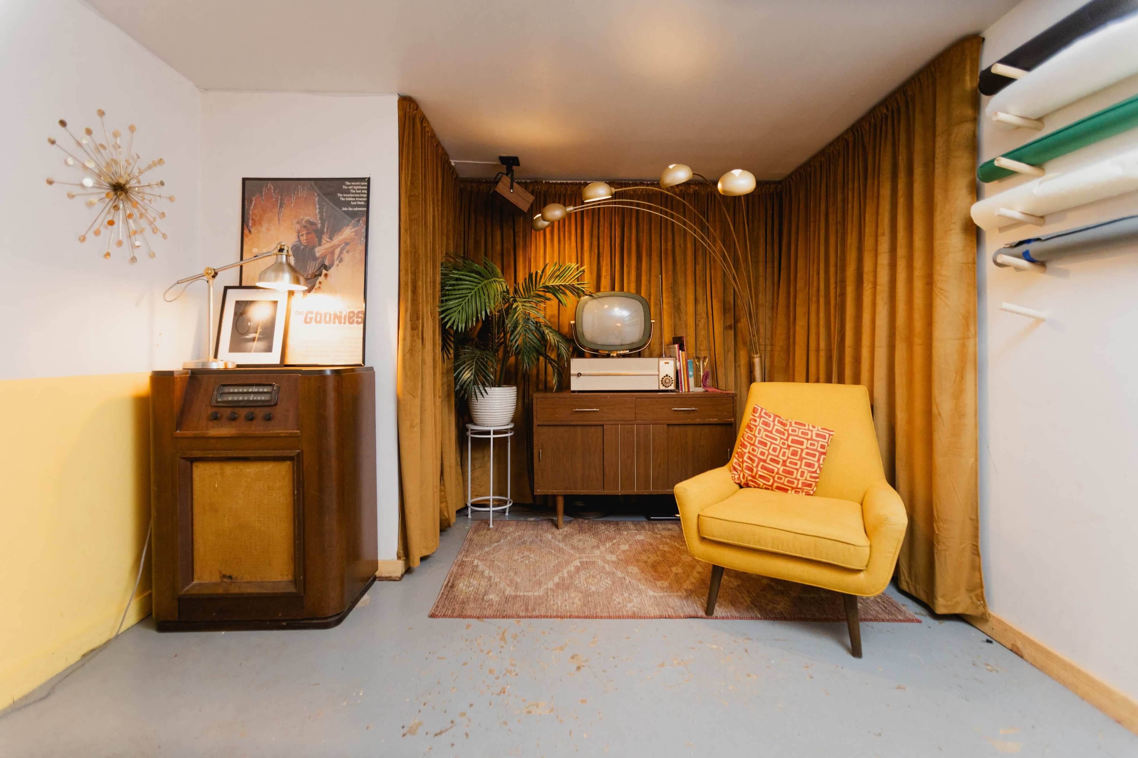 The image shows a retro-styled room featuring a yellow chair, a vintage television, a plant, and a wooden cabinet against a backdrop of yellow and brown curtains.