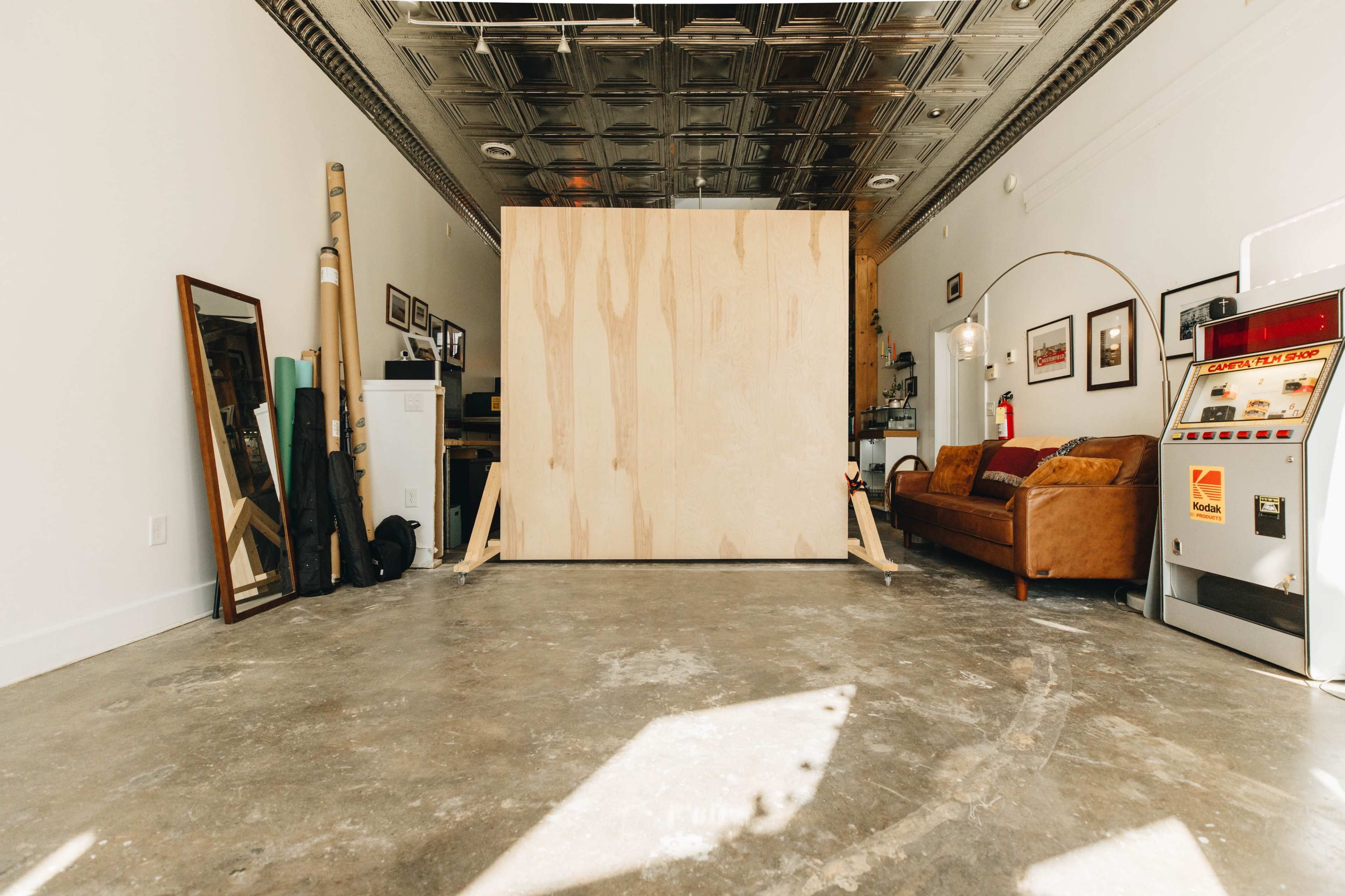 A large plywood panel stands in the center of a spacious room with a concrete floor, flanked by a mirror, a couch, and various items against the walls.