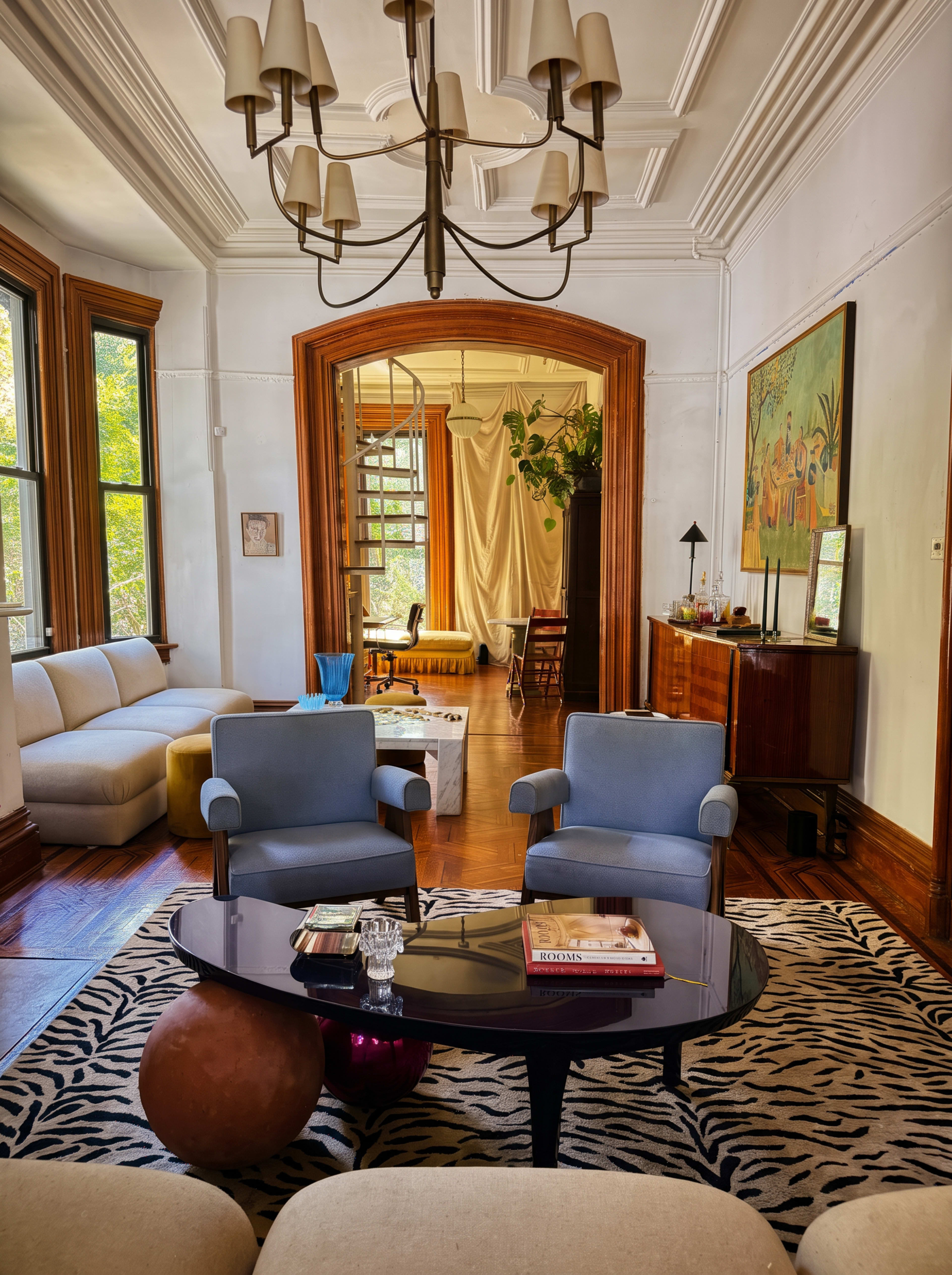 The image shows a stylish living room featuring two blue chairs facing a black coffee table on a patterned rug, with large windows allowing natural light to illuminate the space.