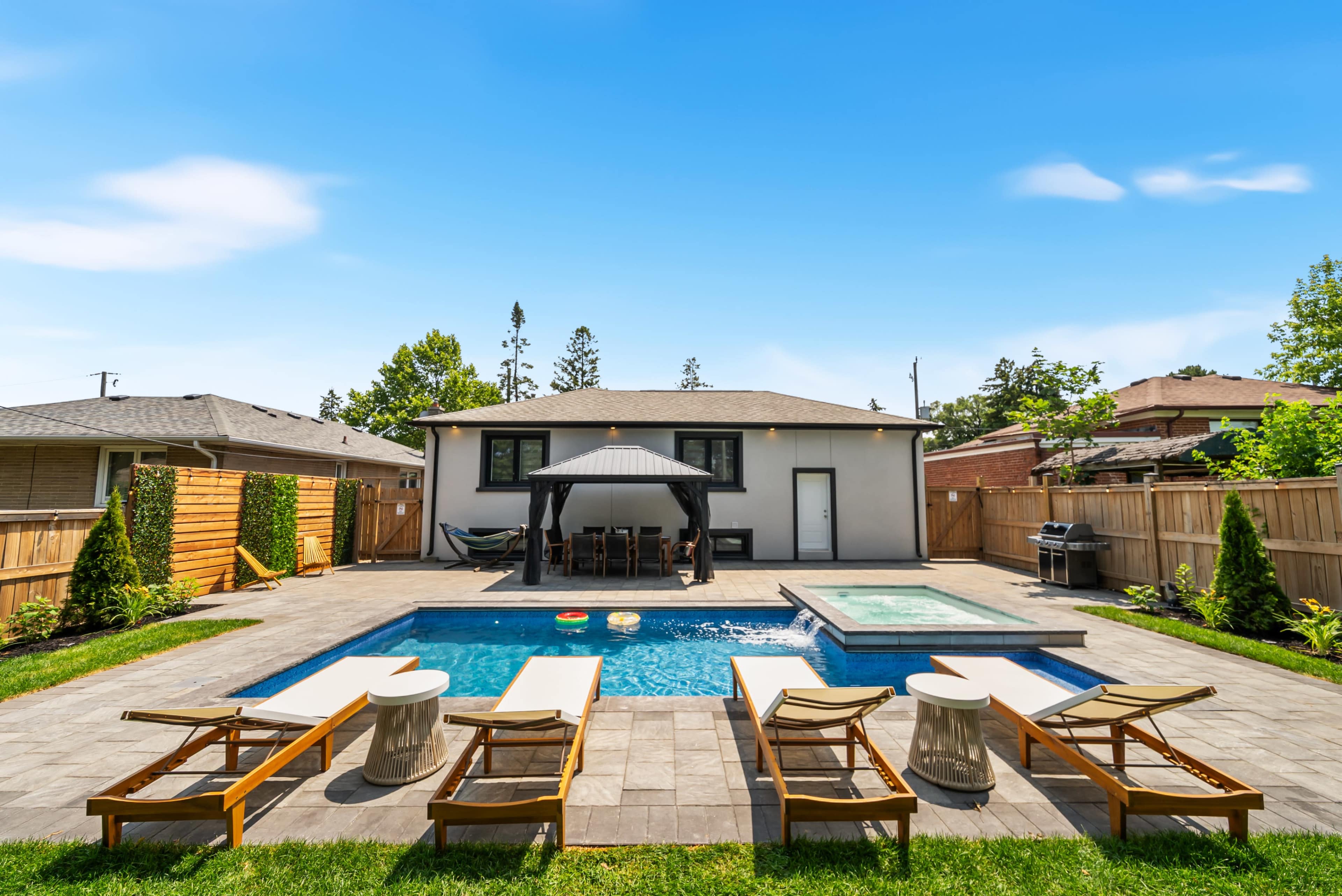 The image shows a backyard with a swimming pool, a hot tub, several lounge chairs, and a patio area near a modern house.