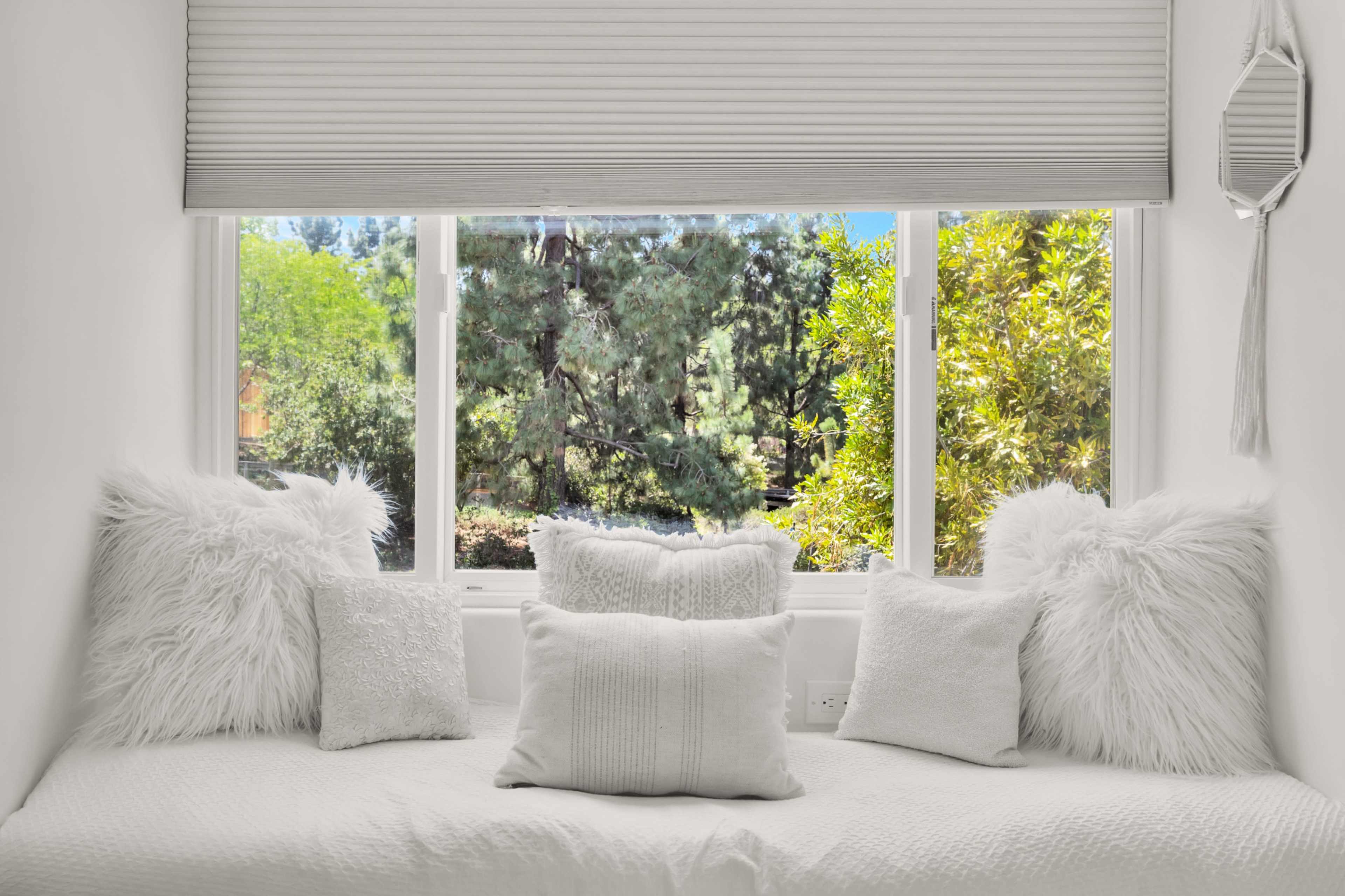 A cozy window nook with white pillows and throws overlooks a vibrant garden.