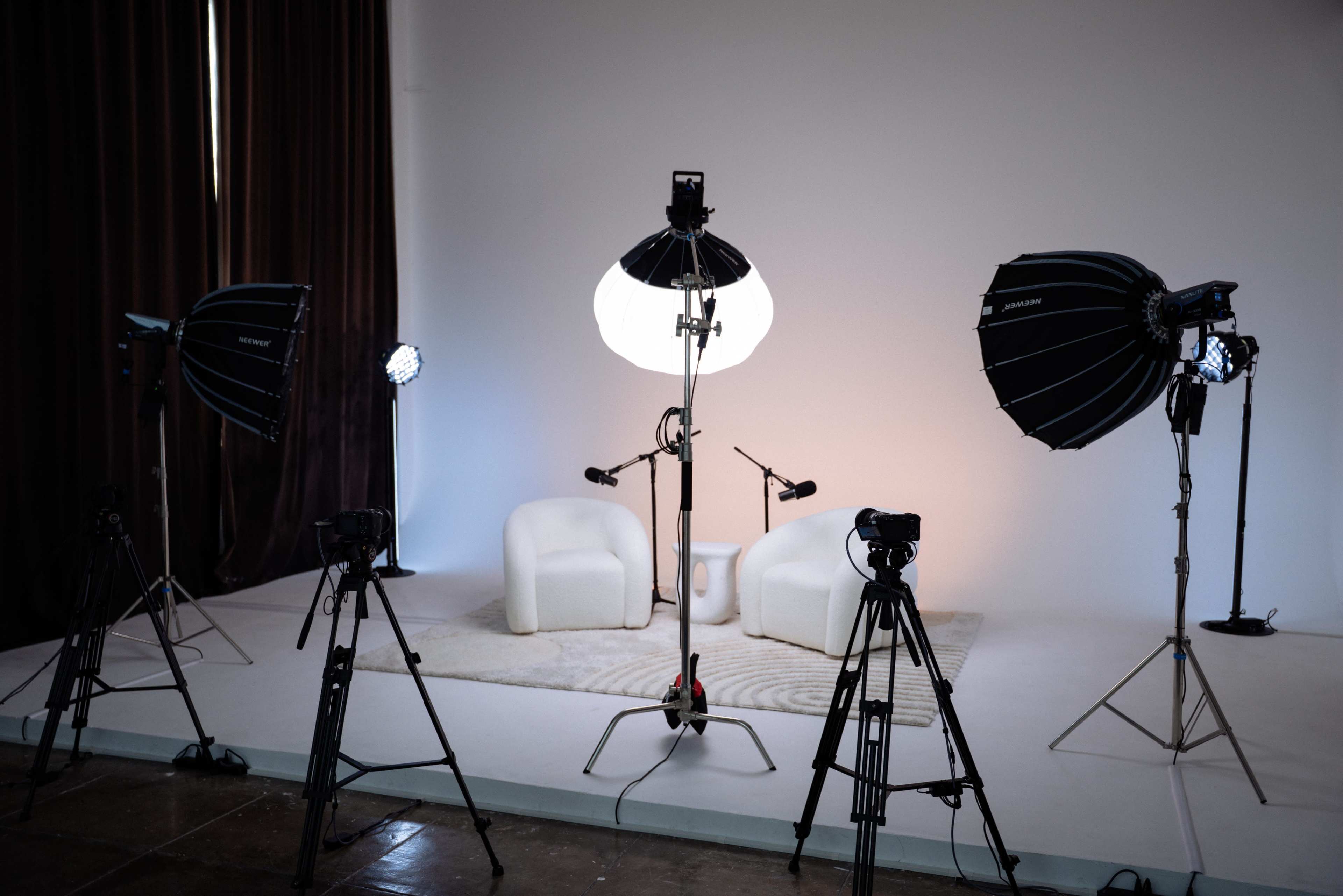 The image shows a photography studio setup with two white chairs, three camera tripods, and multiple light sources against a blank backdrop.