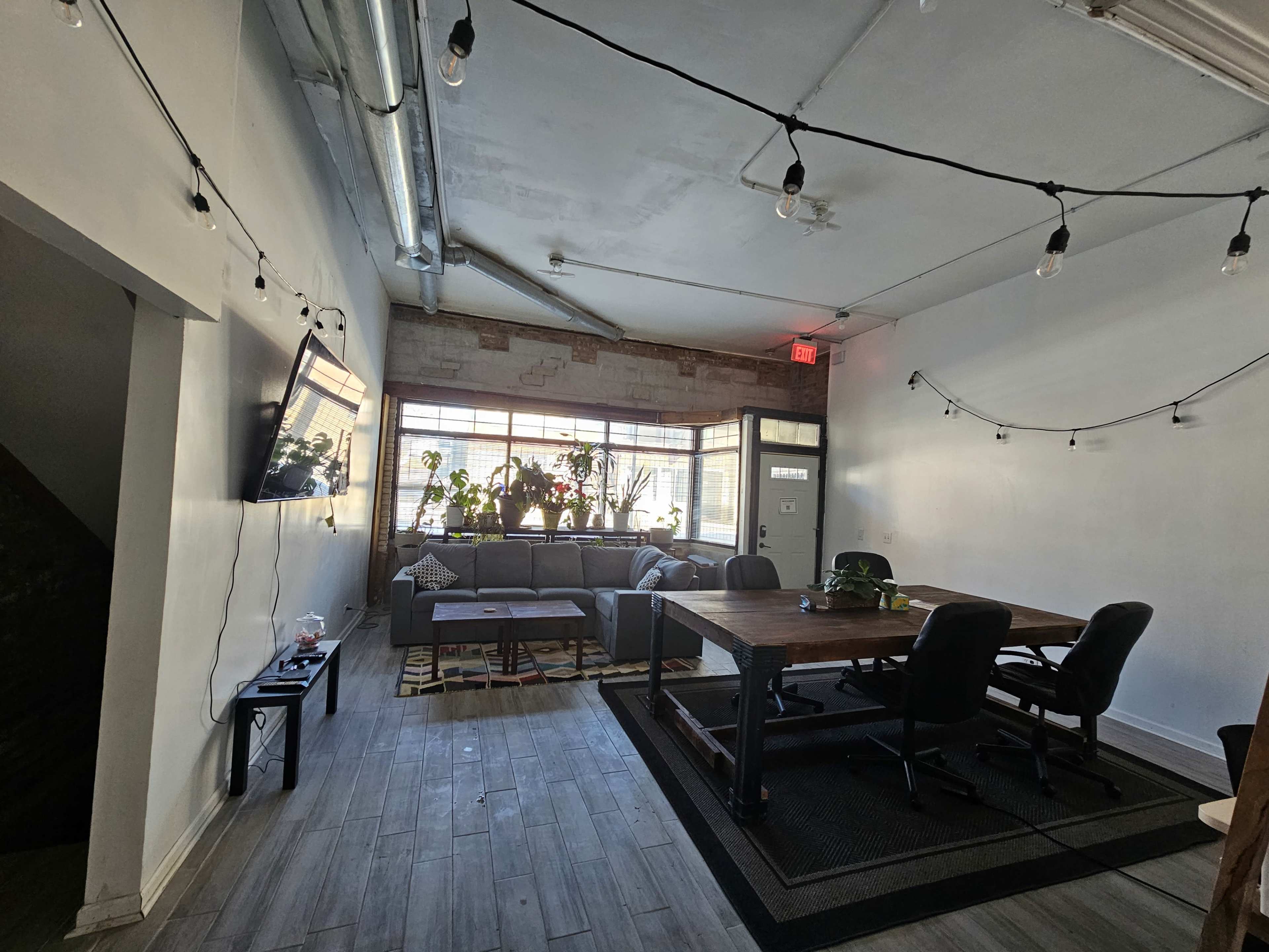 The image shows a modern office space with a seating area featuring a sofa and plants, alongside a large wooden table with chairs.