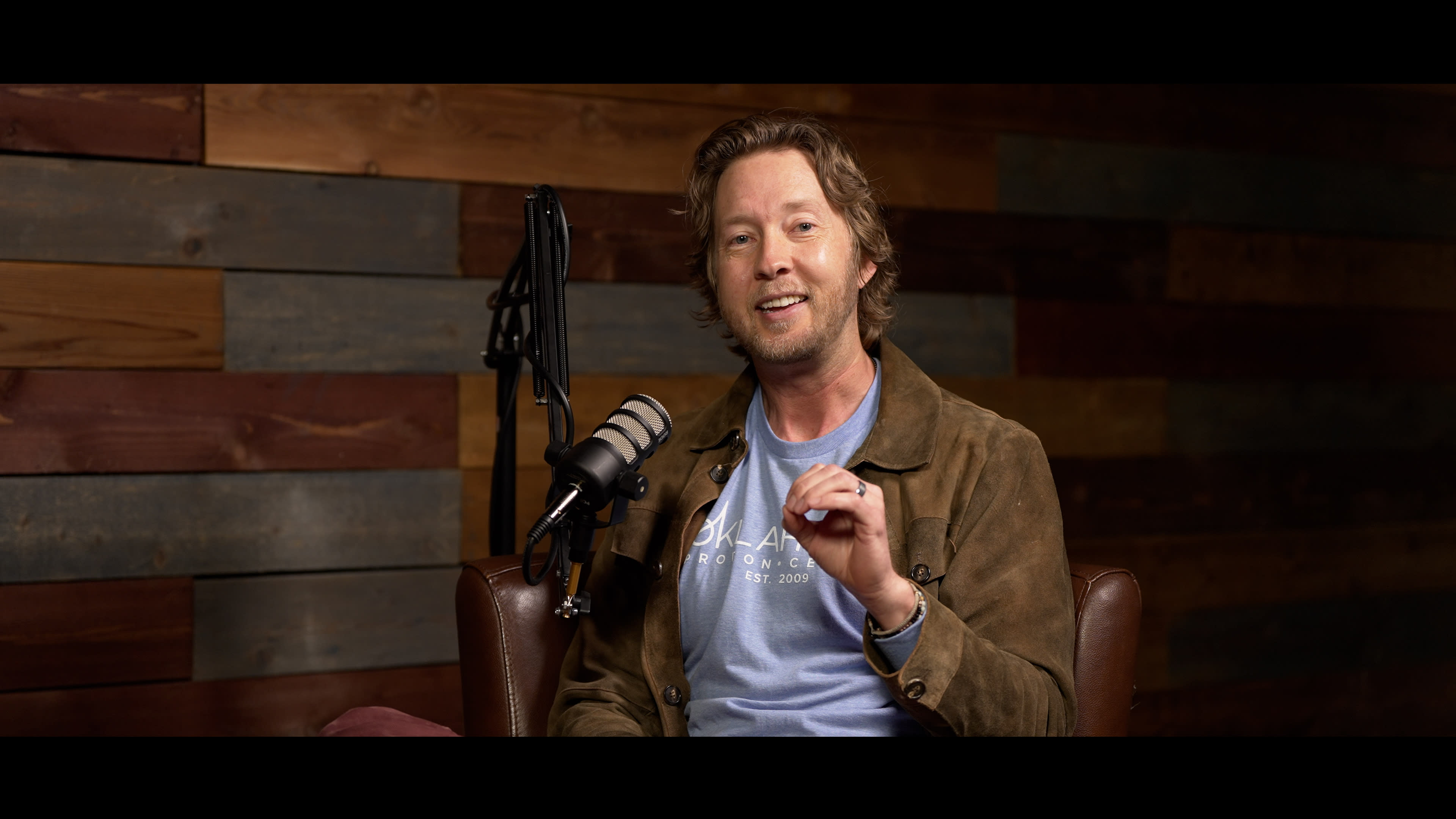 A man is seated on a brown chair in front of a microphone, smiling while gesturing with his hand against a backdrop of wooden panels.