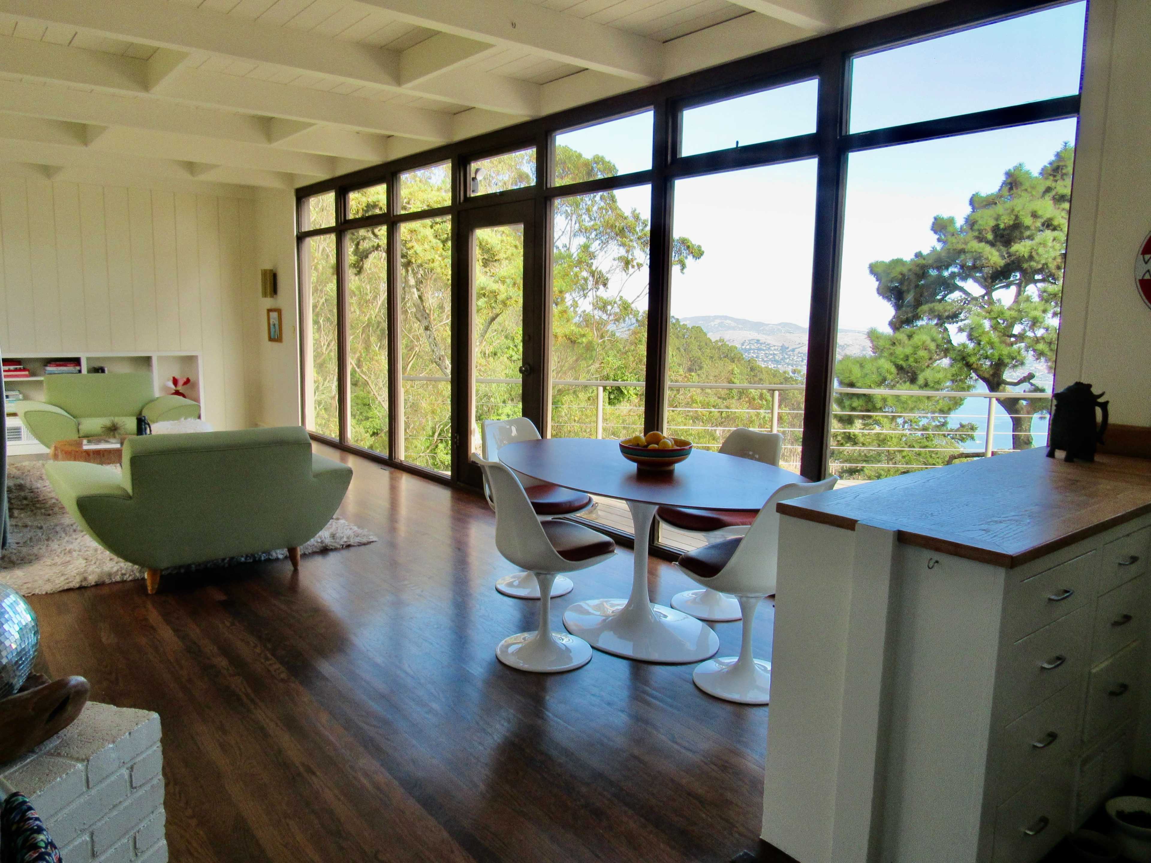 A bright, modern living space with large windows offering a view of trees and a distant landscape, furnished with a green sofa, a dining table, and white chairs.