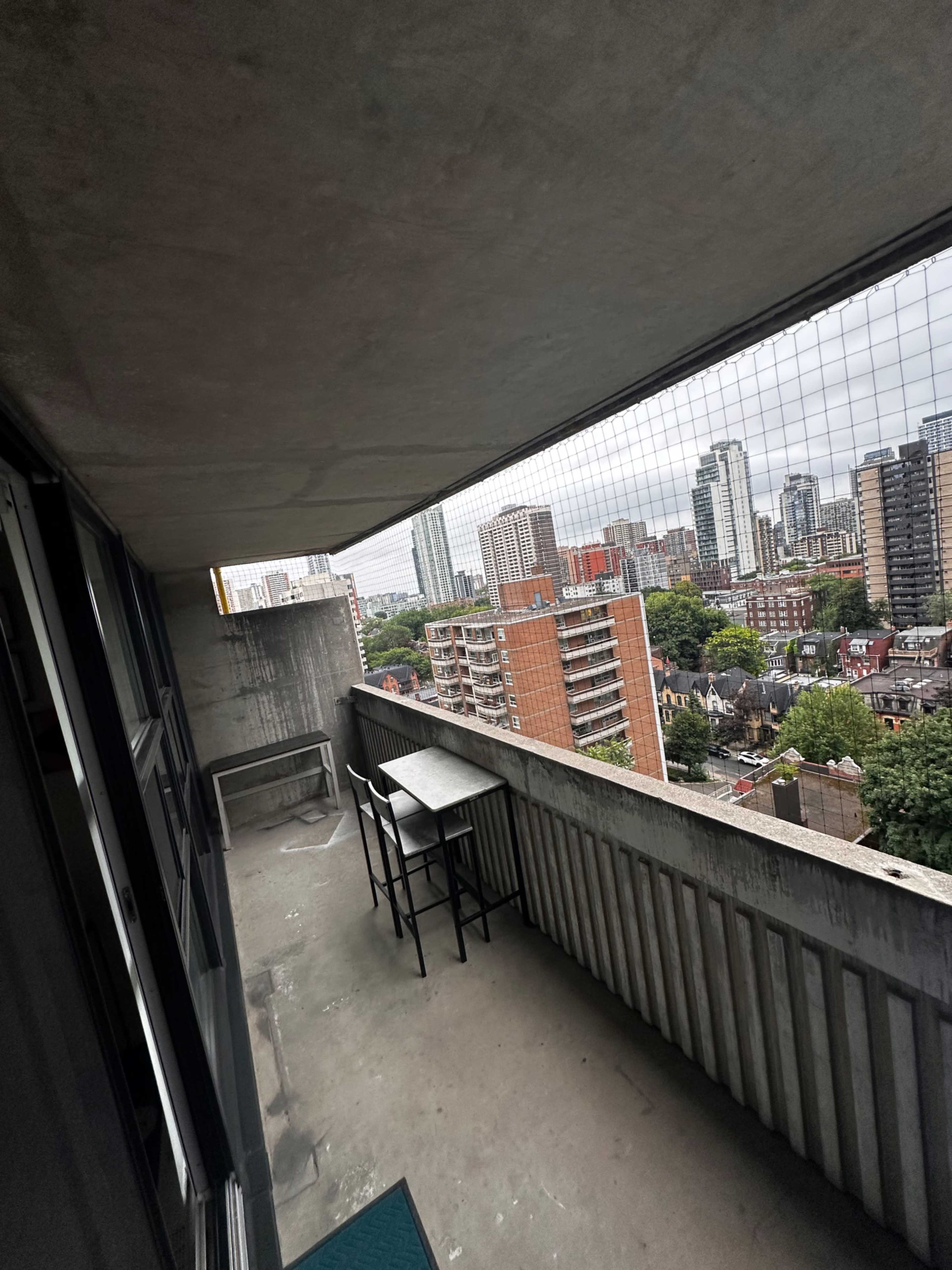 A concrete balcony with a small table and chairs overlooks a cityscape filled with buildings and greenery under a cloudy sky.